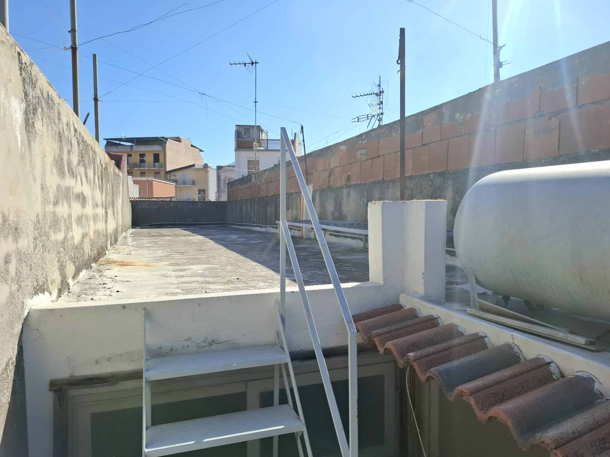 Escaletta in metallo con accesso a un seminterrato, situata su un terrazzo urbano con vista su edifici e linee elettriche, sotto un cielo sereno.