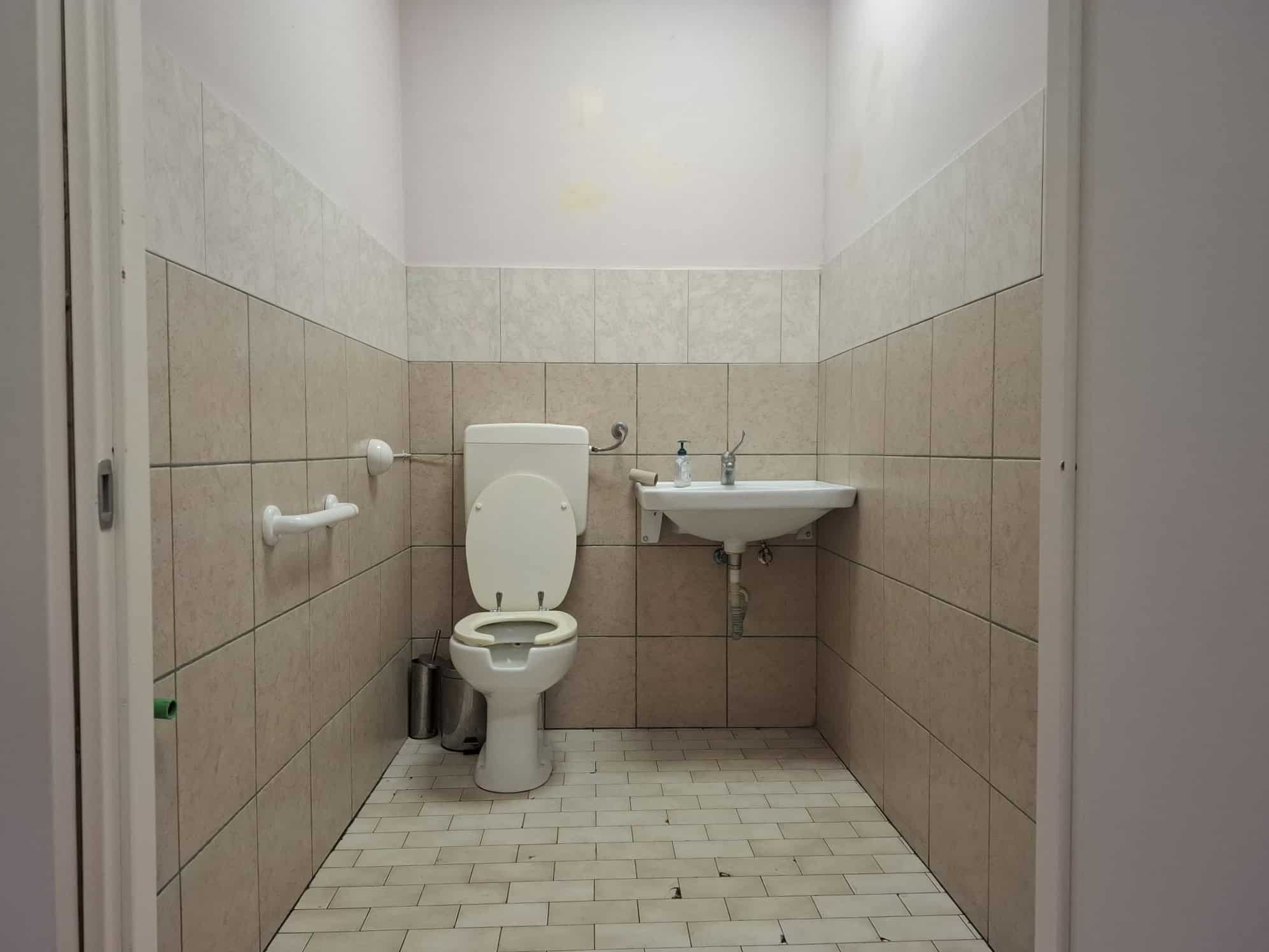 Bagno pubblico con WC e lavabo, ambiente semplice e funzionale, ideale per strutture pubbliche o uffici, con pareti rivestite in piastrelle beige e pavimento in piastrelle chiare.