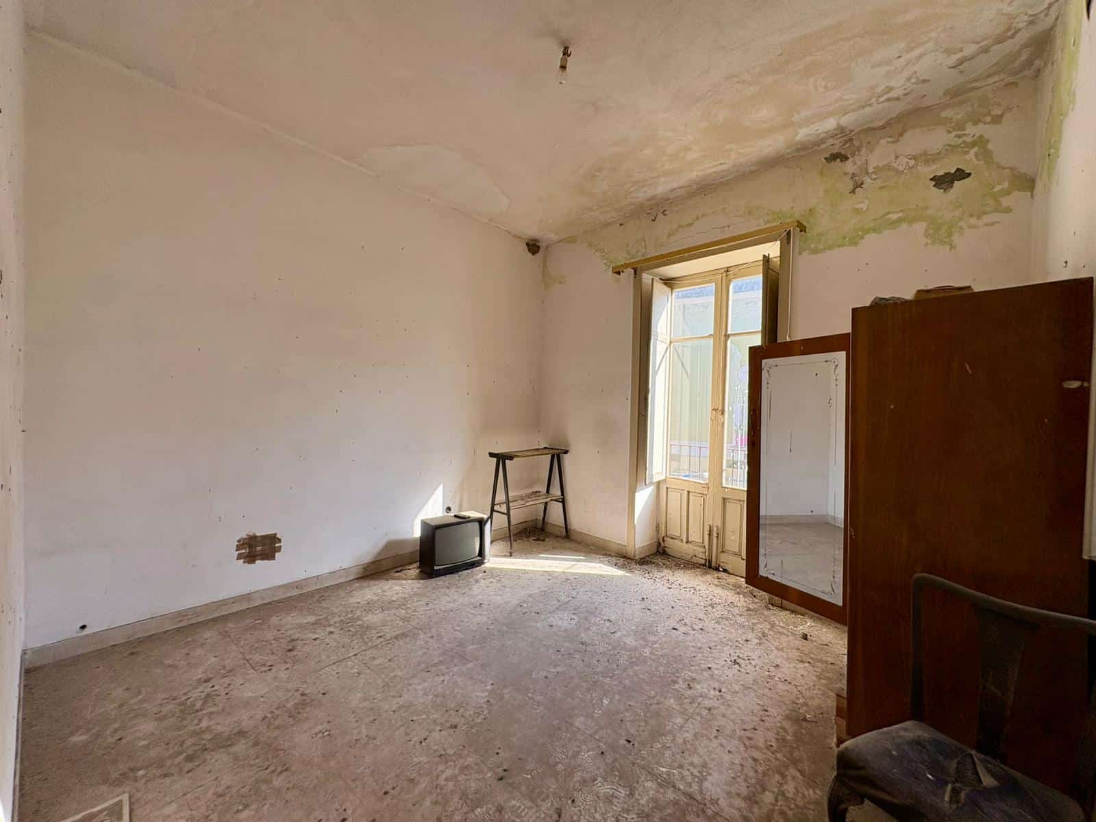 Una stanza vuota e trascurata con pareti e soffitto danneggiati, pavimento in cemento e una finestra che lascia entrare luce naturale, con mobili vecchi e deteriorati.
