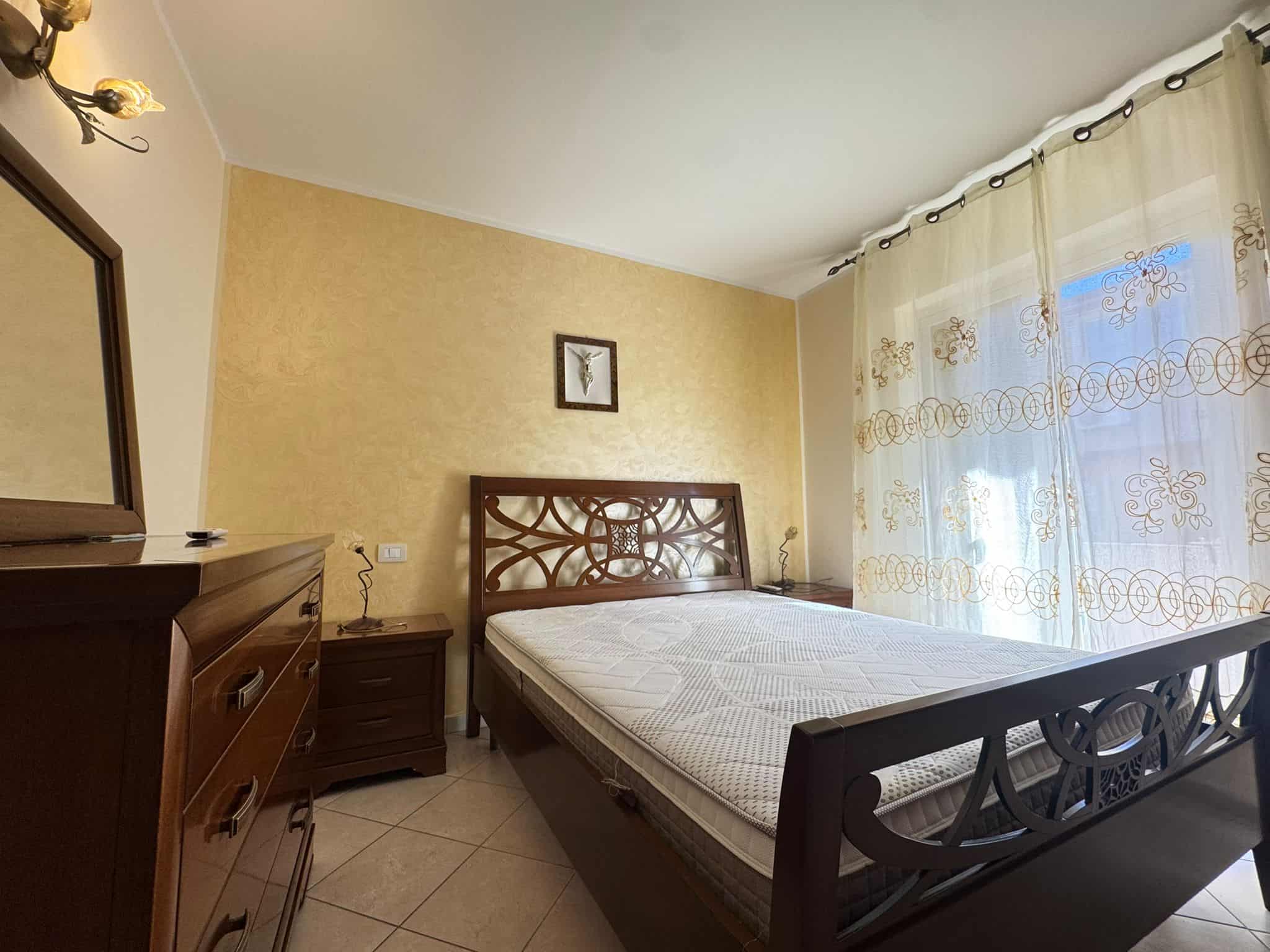 Camera da letto luminosa con pareti beige, letto in legno, armadio e finestra con tende decorative, ambiente accogliente e ben illuminato.