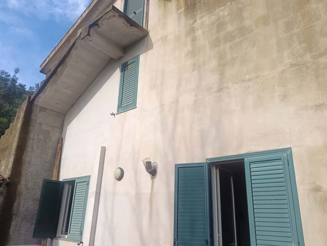 Ampia facciata di casa con pareti in stucco chiaro, finestre con persiane blu e un'illuminazione esterna, situata in un contesto residenziale soleggiato.