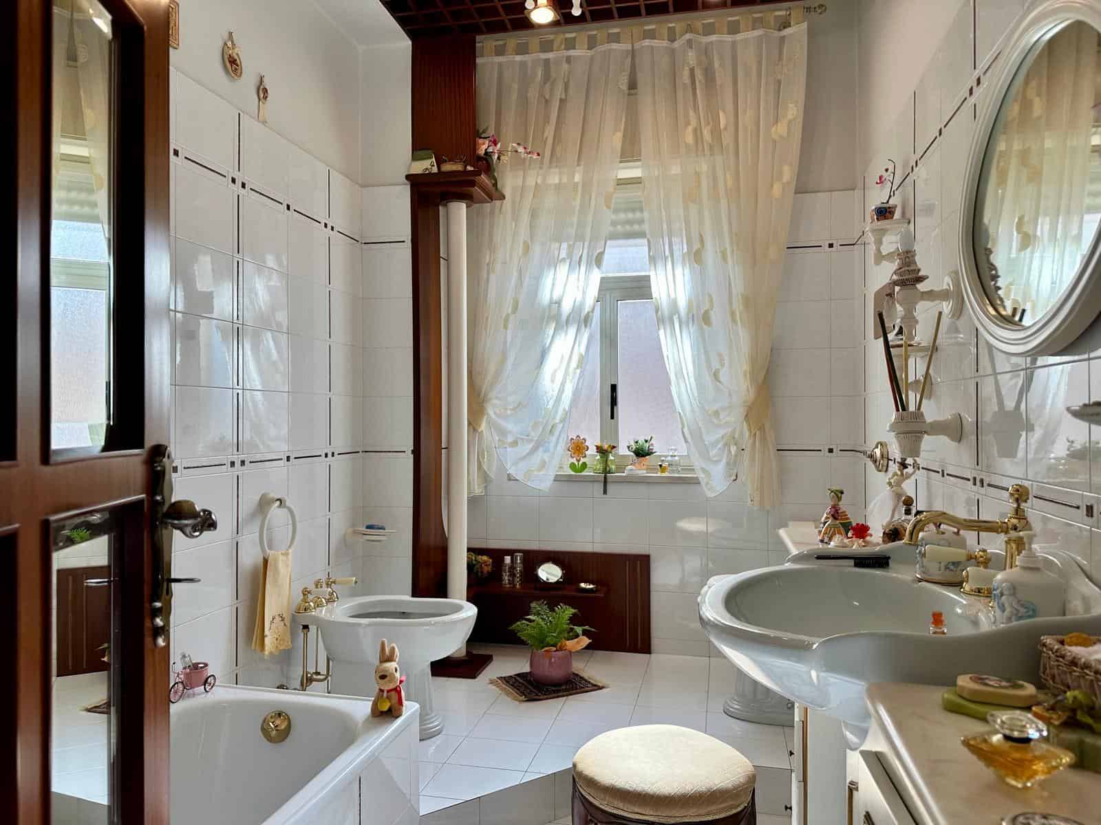 Doccia e lavabo in un bagno elegante con dettagli classici, illuminato da luce naturale attraverso una finestra con tende leggere, decorazioni floreali e accessori raffinati.