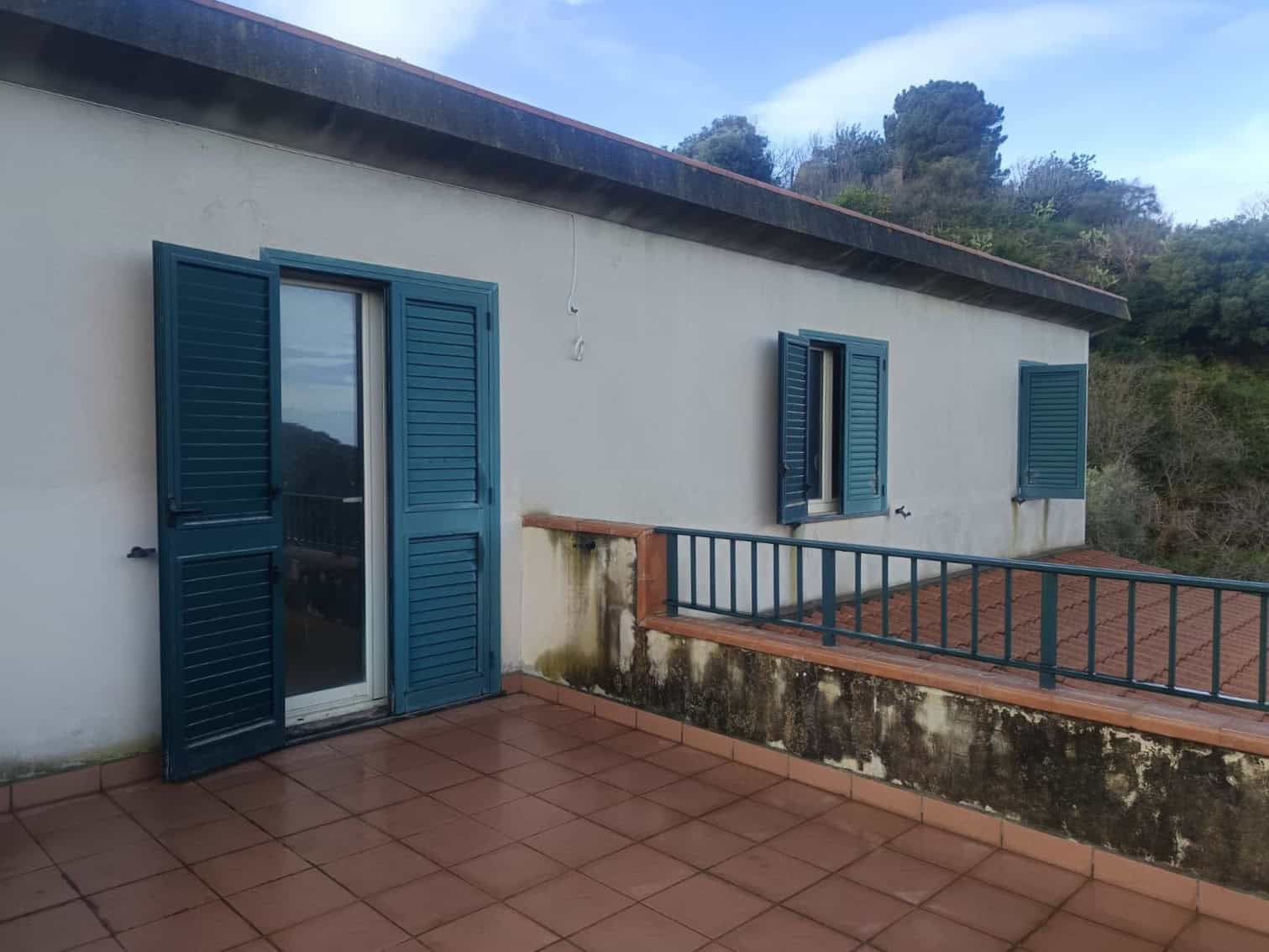 Balcone con persiane blu e vista su natura, ideale per relax e momenti all'aperto in una casa in stile mediterraneo. Spazio esterno con pavimentazione in terracotta e vista panoramica su colline verdi.