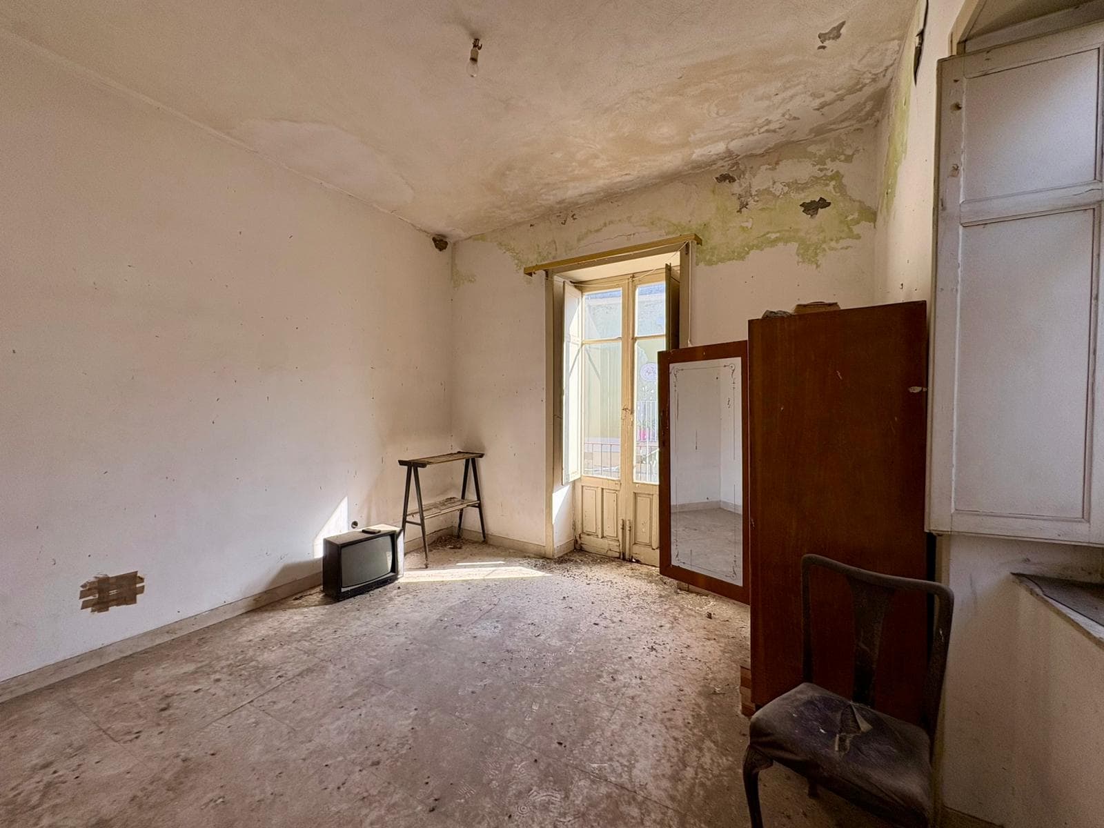Vecchio ambiente interno di una casa con pareti danneggiate e soffitto usurato, finestra con vista su un balcone, mobili vecchi e un televisore anni '80, atmosfera di abbandono e decadenza.