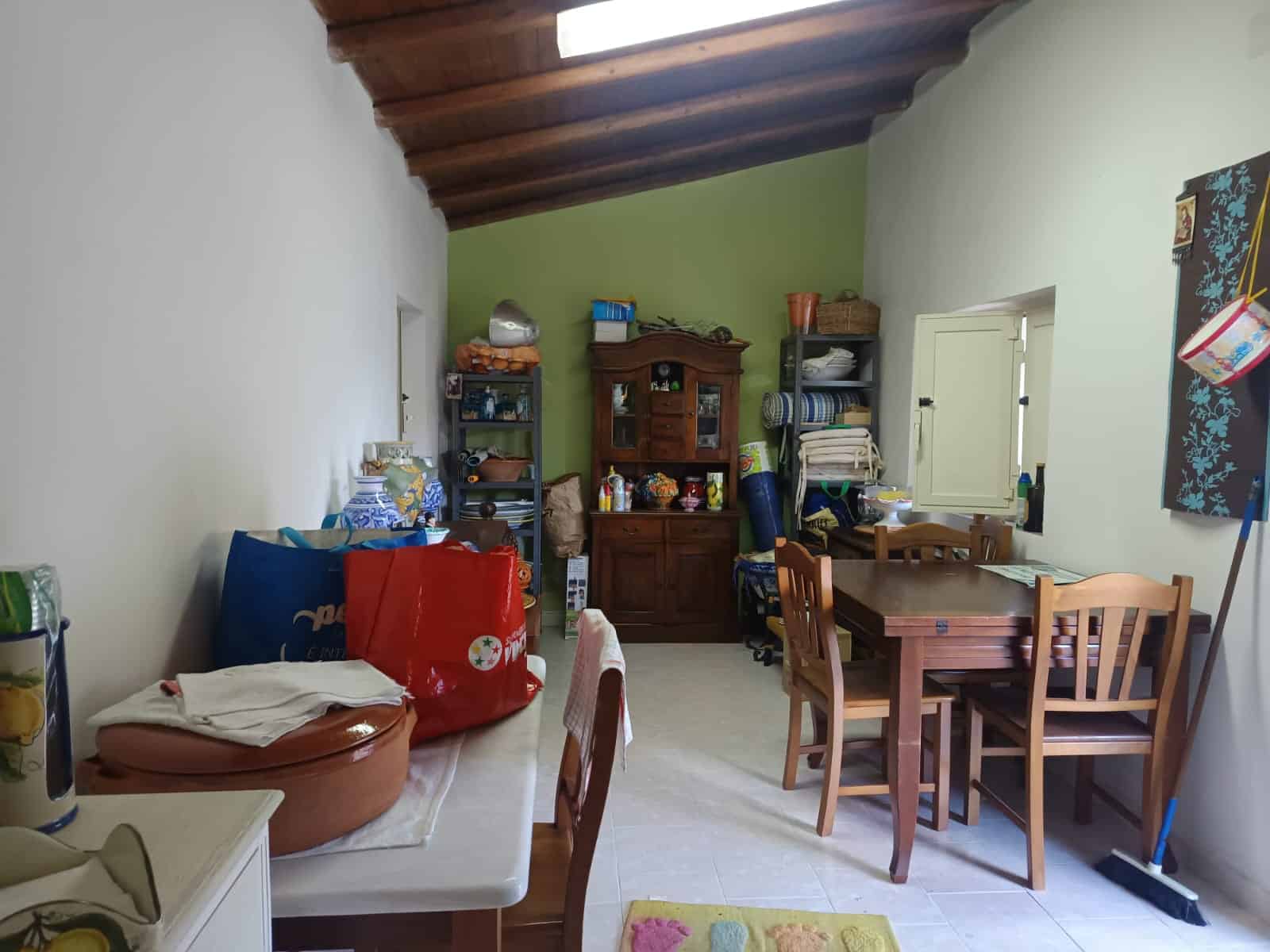 Ampia stanza con soffitto in legno, arredi in legno, tavolo da pranzo con sedie, scaffali con oggetti vari, finestra che lascia entrare luce naturale, ambiente accogliente e funzionale.