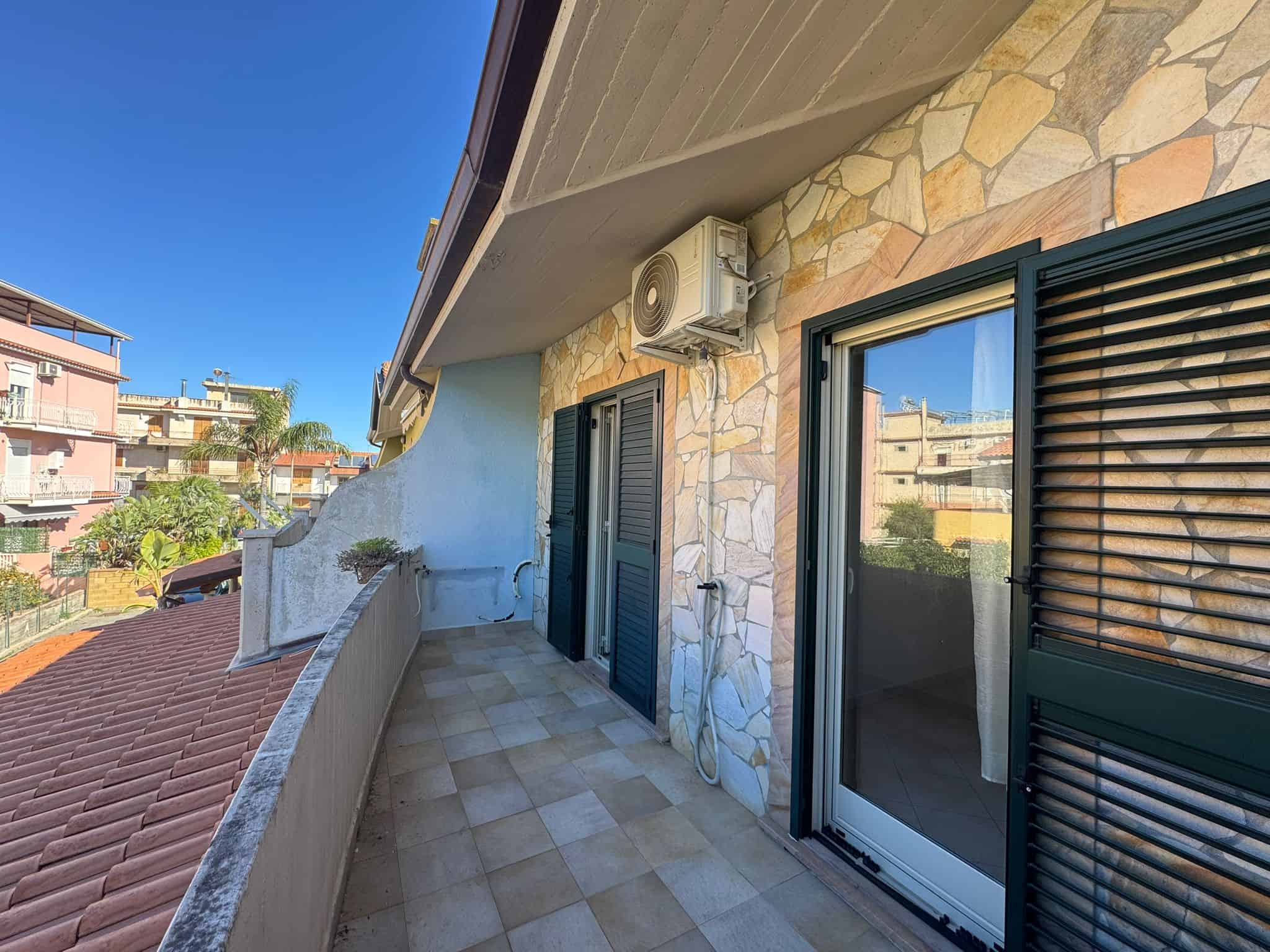 Balcone di un appartamento con vista sulla città, rivestito in pietra e dotato di aria condizionata, con ampie finestre e porte scorrevoli in vetro, ideale per relax e svago all'aperto.