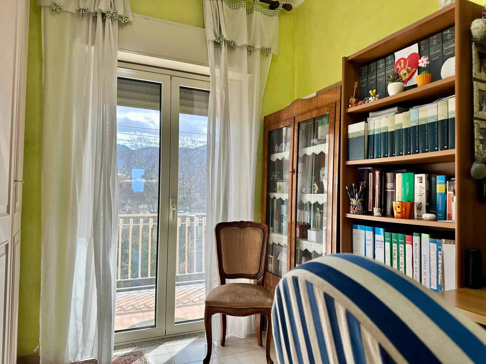 Interno di una stanza luminosa con libreria, sedia e porta finestra che dà su un balcone con vista sulle montagne.