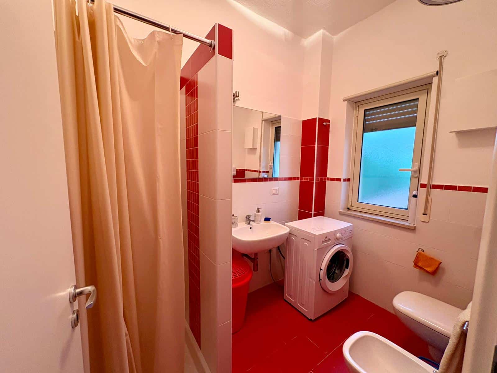 Doccia e lavatrice in bagno con pareti bianche e dettagli rossi, finestra con tapparella, arredo funzionale e spazio ottimizzato per il comfort quotidiano.