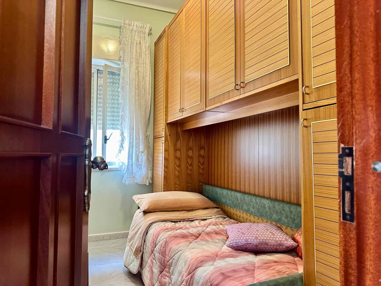 Camera da letto con armadiature in legno, letto con cuscini e tende leggere, ambiente luminoso e accogliente, ideale per case in vendita o affitto.