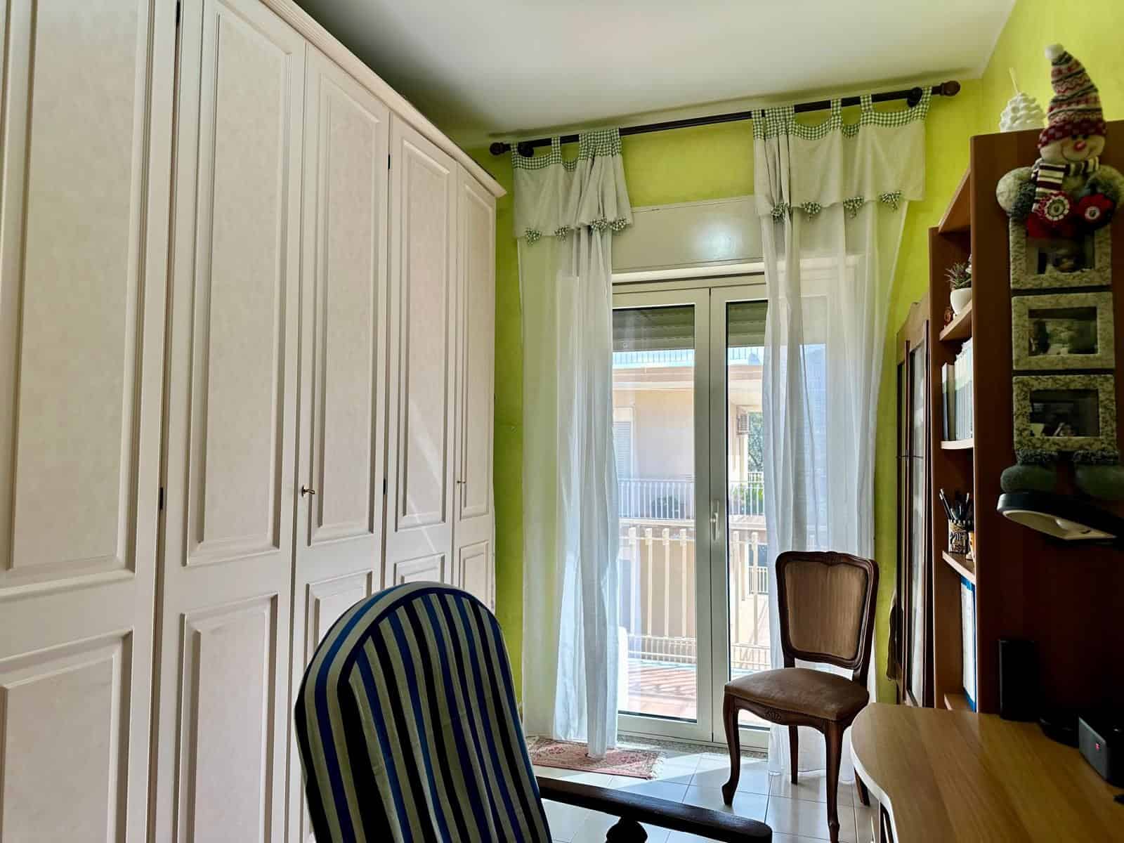 Una stanza luminosa con pareti verde lime, armadio bianco, e una porta finestra che dà su un balcone, decorata con tende leggere e trasparenti.