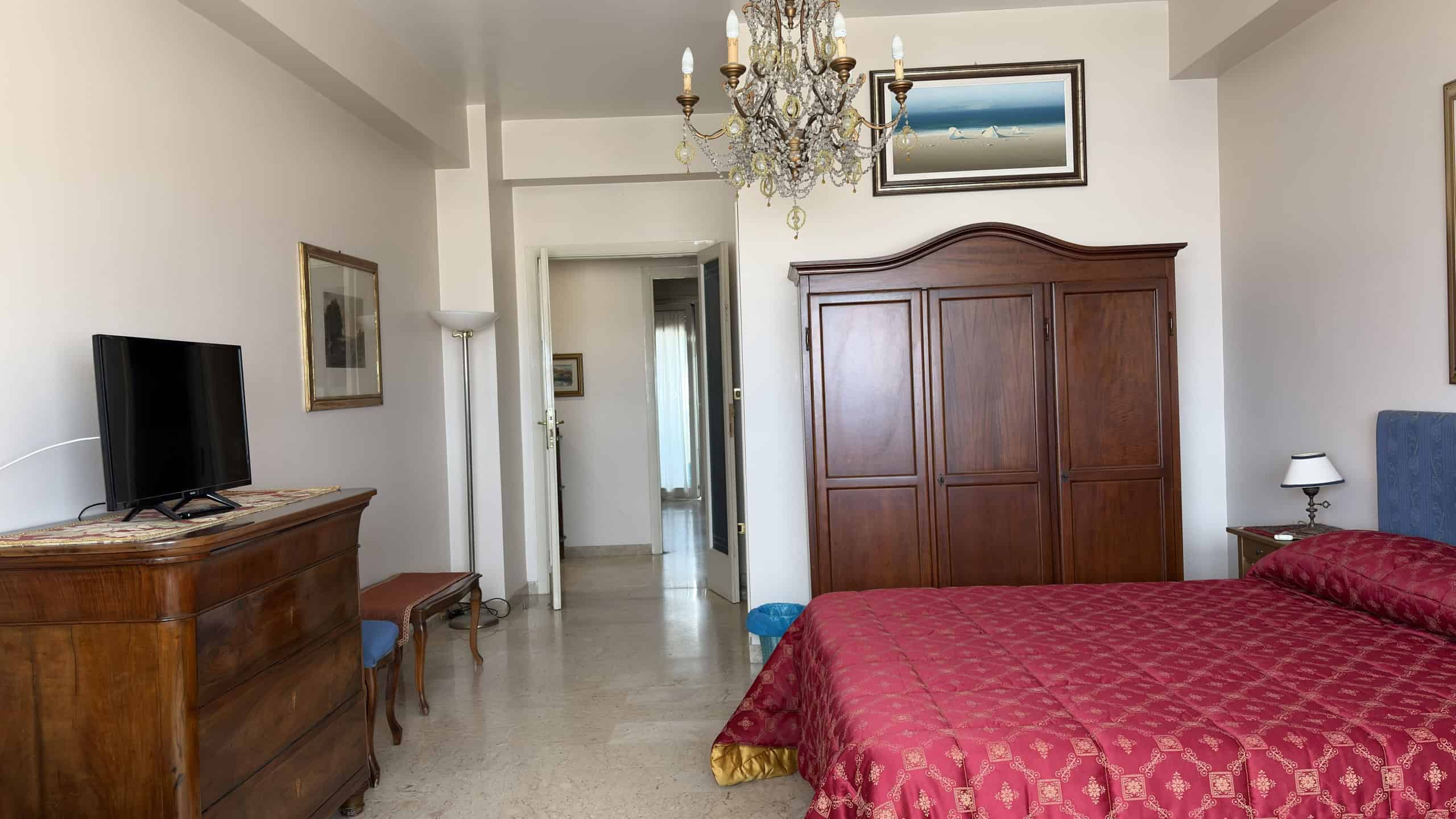 Elegante camera da letto con arredi classici, grande armadio in legno e letto con copriletto rosso, illuminata da lampade e lampadari, ideale per soggiorni confortevoli e raffinati.