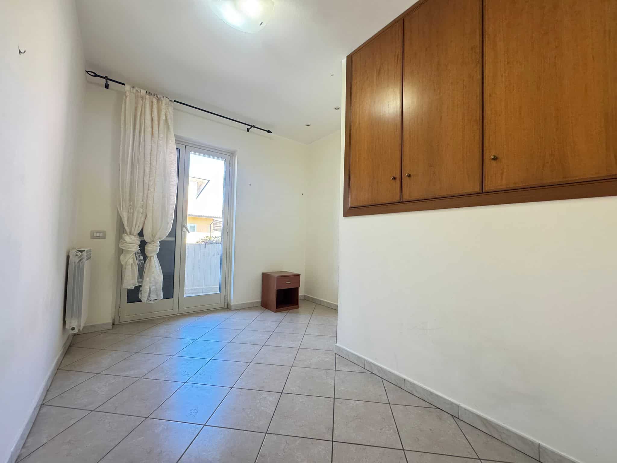 Aggiungi un'immagine di una stanza vuota con pareti bianche, un grande armadio in legno, una porta finestra con tende leggere e un piccolo mobile in legno, ideale per annunci immobiliari e affitti.