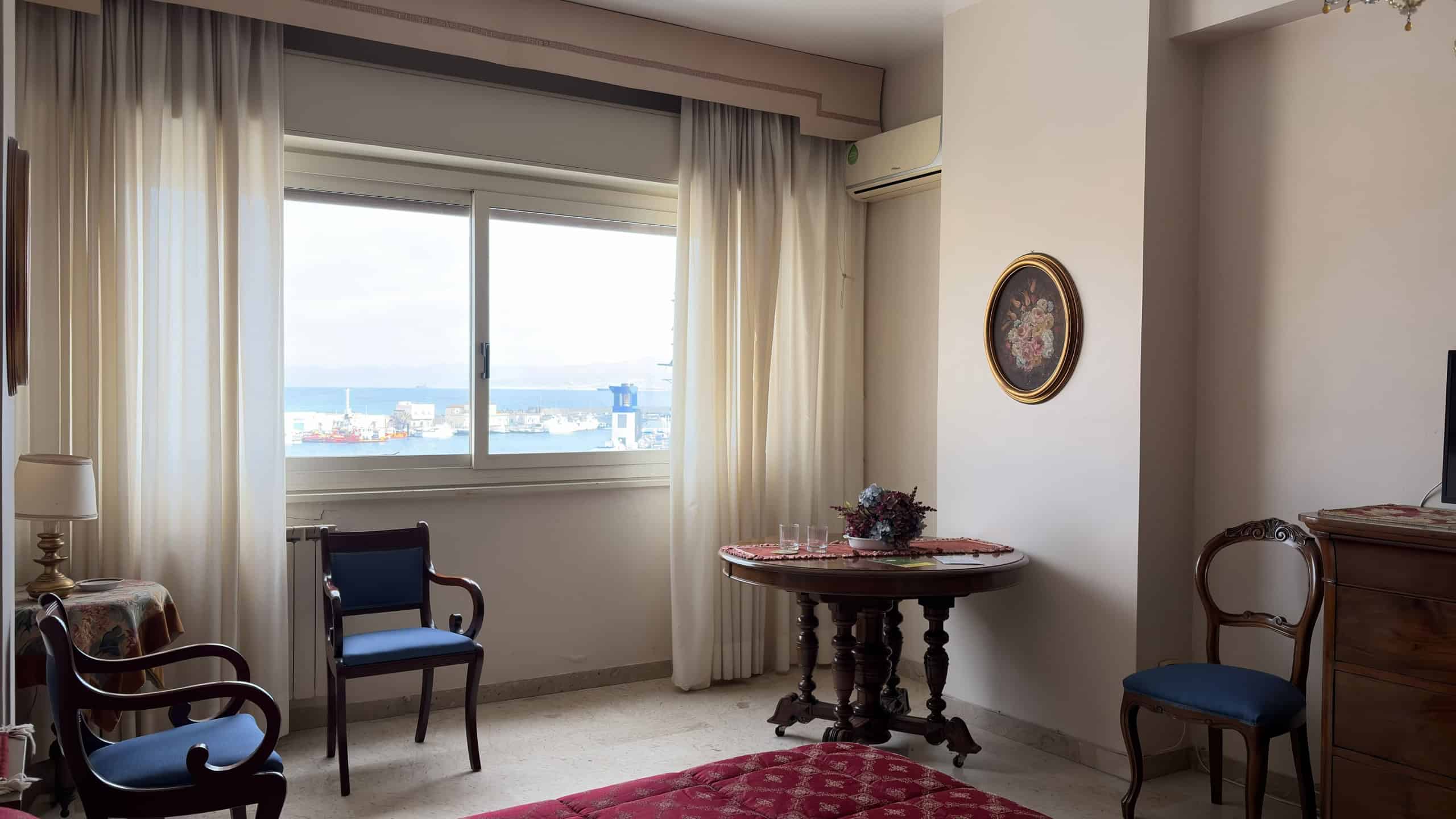 Vista panoramica di un soggiorno luminoso con grandi finestre che offrono una vista sul mare e sulla città, arredato con sedie in legno e un tavolo centrale, decorazioni eleganti e tende leggere.