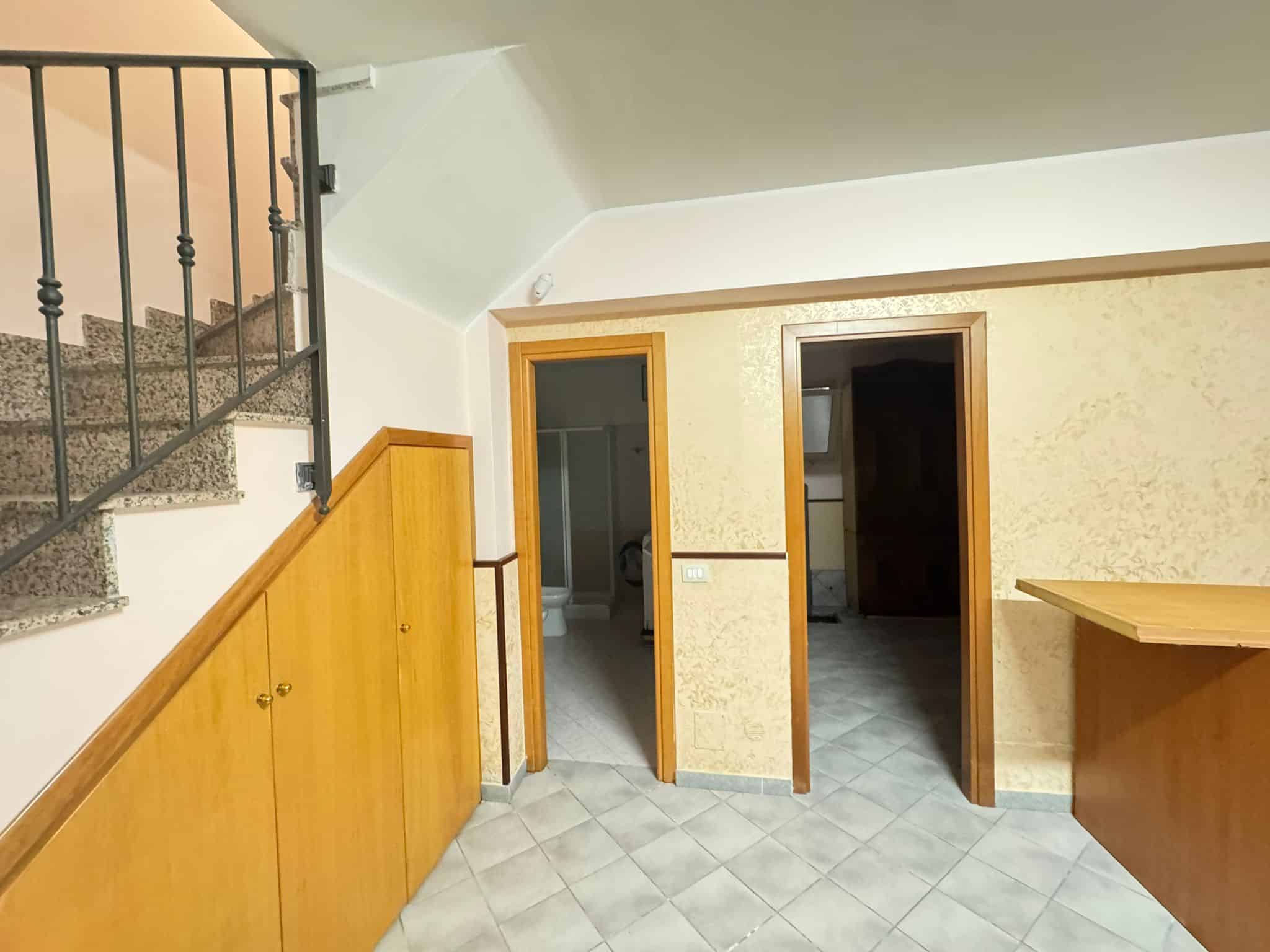 Ampia zona giorno con pareti gialle e pavimento in piastrelle, vista su due porte che conducono a stanze diverse, con arredi in legno e una scala in ferro e pietra che sale verso l'alto.