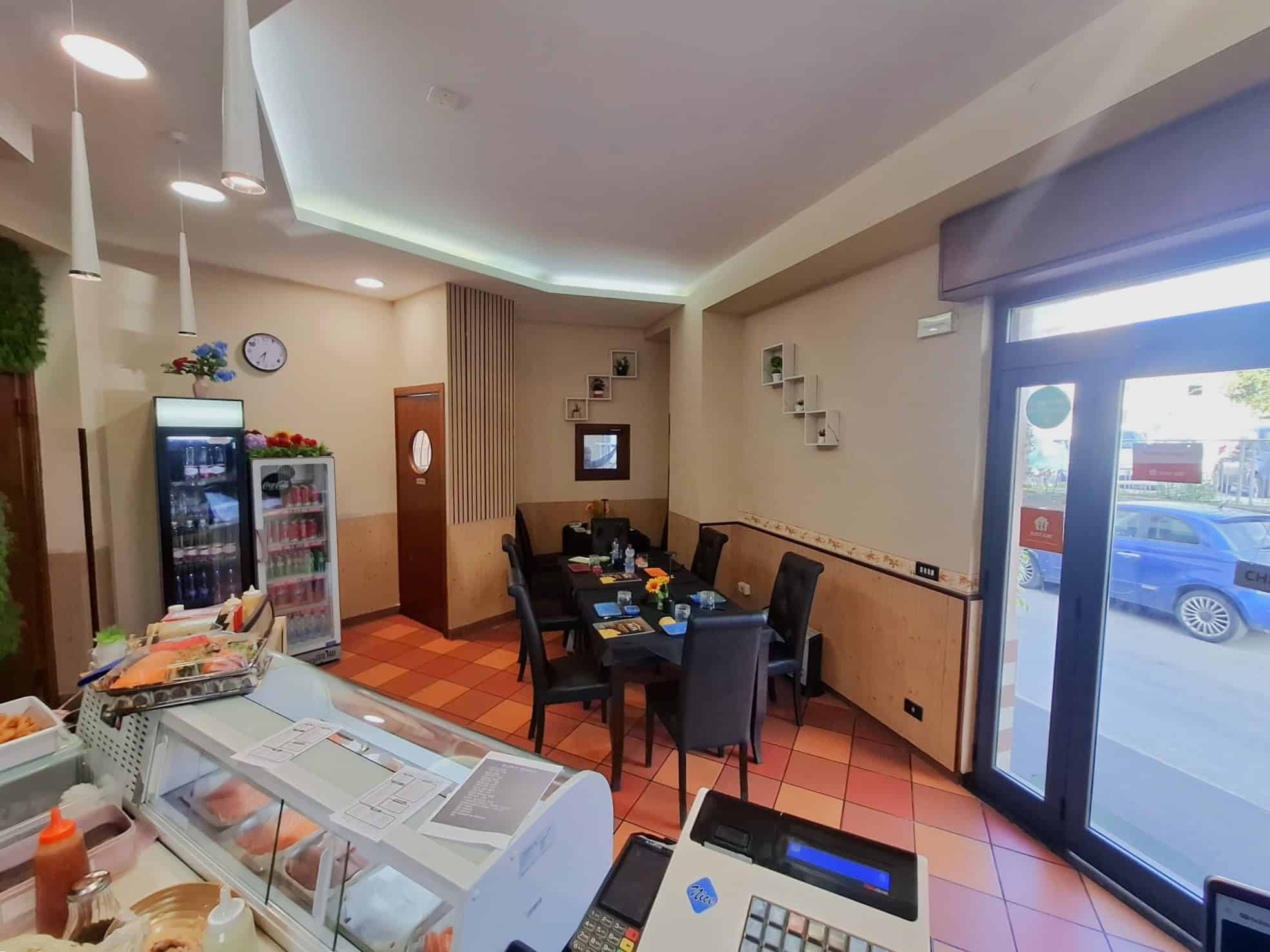 Primo piano di un ristorante o caffetteria con tavoli apparecchiati, area di servizio con frigorifero e banco alimentare, ambiente accogliente e moderno con illuminazione soffusa e decorazioni elegant.