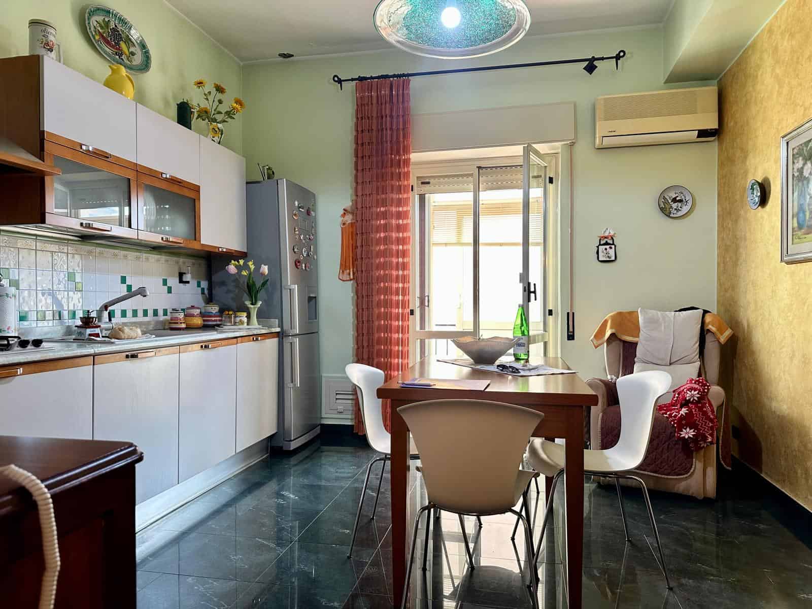 Una cucina luminosa e accogliente con pareti verde pastello, mobili bianchi, e un tavolo centrale con sedie, ideale per vivere momenti conviviali in un ambiente domestico confortevole.