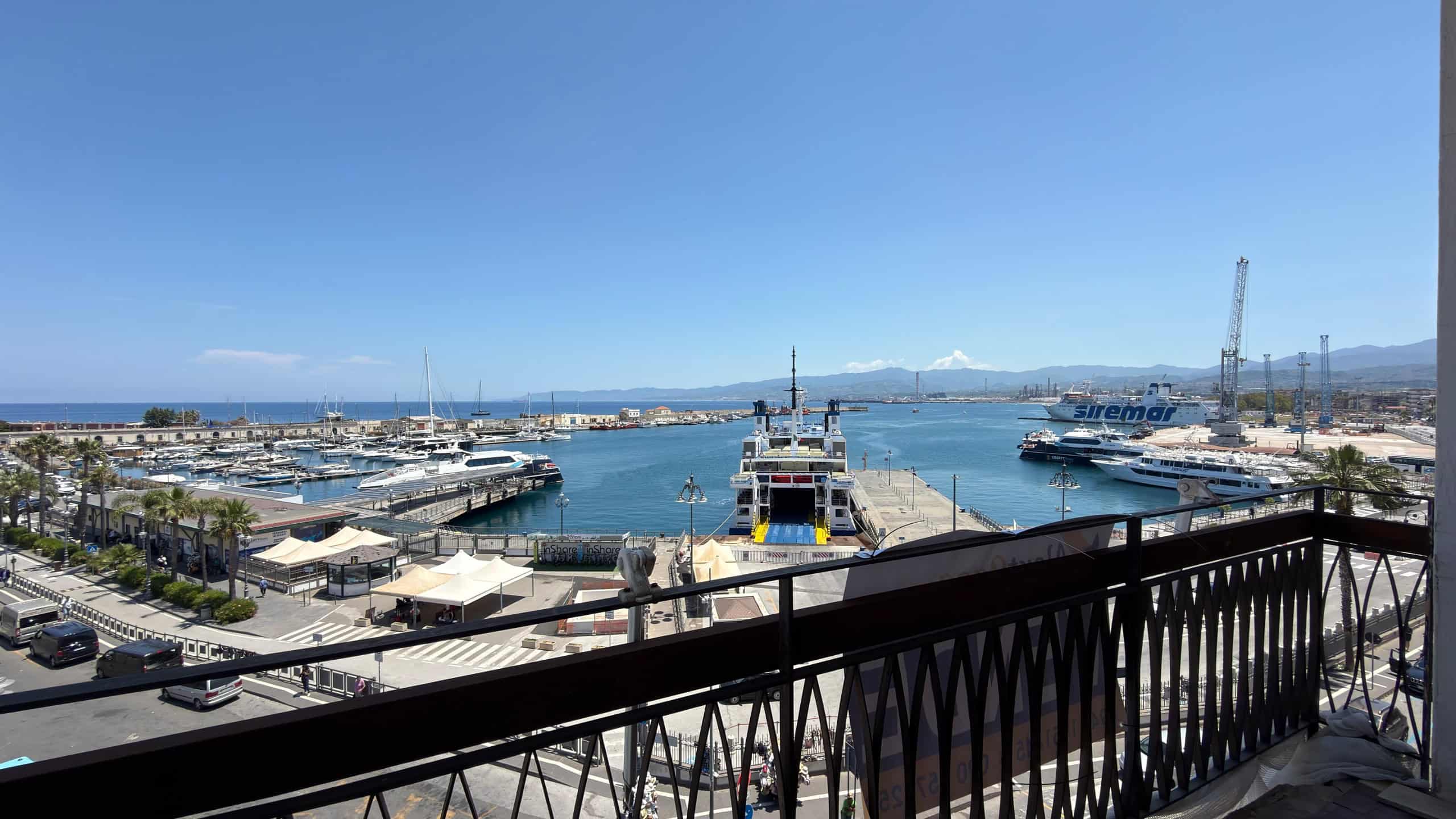 Vista panoramica del porto di mare con barche e yacht, vista dal balcone di un appartamento a NextCasa, in una giornata soleggiata con cielo azzurro e montagne sullo sfondo.
