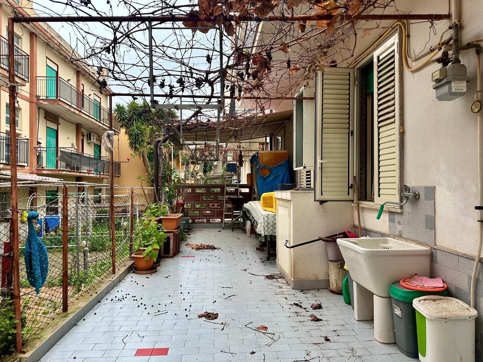 Balcone di un appartamento con piante, arredi e tettoia in legno, vista su cortile urbano, in stile abitativo mediterraneo.