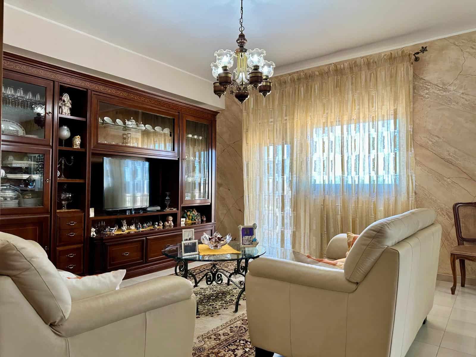Elegante soggiorno con mobili in legno scuro, divani in pelle beige, grande finestra con tende leggere, lampadario antico e decorazioni classiche, ideale per vivere momenti di relax e convivialità.