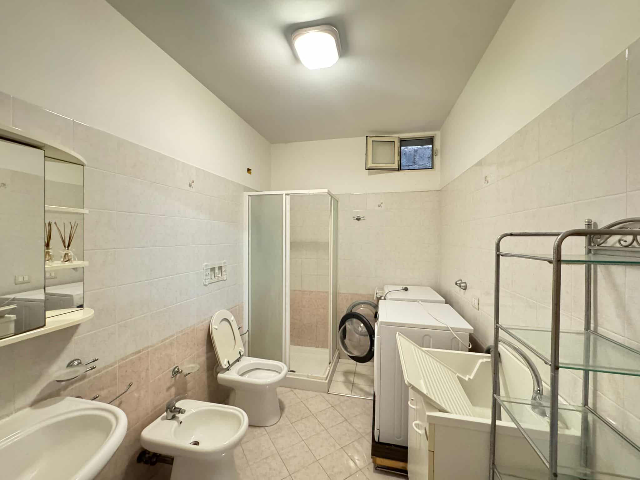 Bagno con sanitari e lavatrice, illuminato da luce artificiale e finestra piccola, con pareti rivestite in piastrelle chiare e pavimento in piastrelle beige, in un ambiente funzionale e ben organizzat.