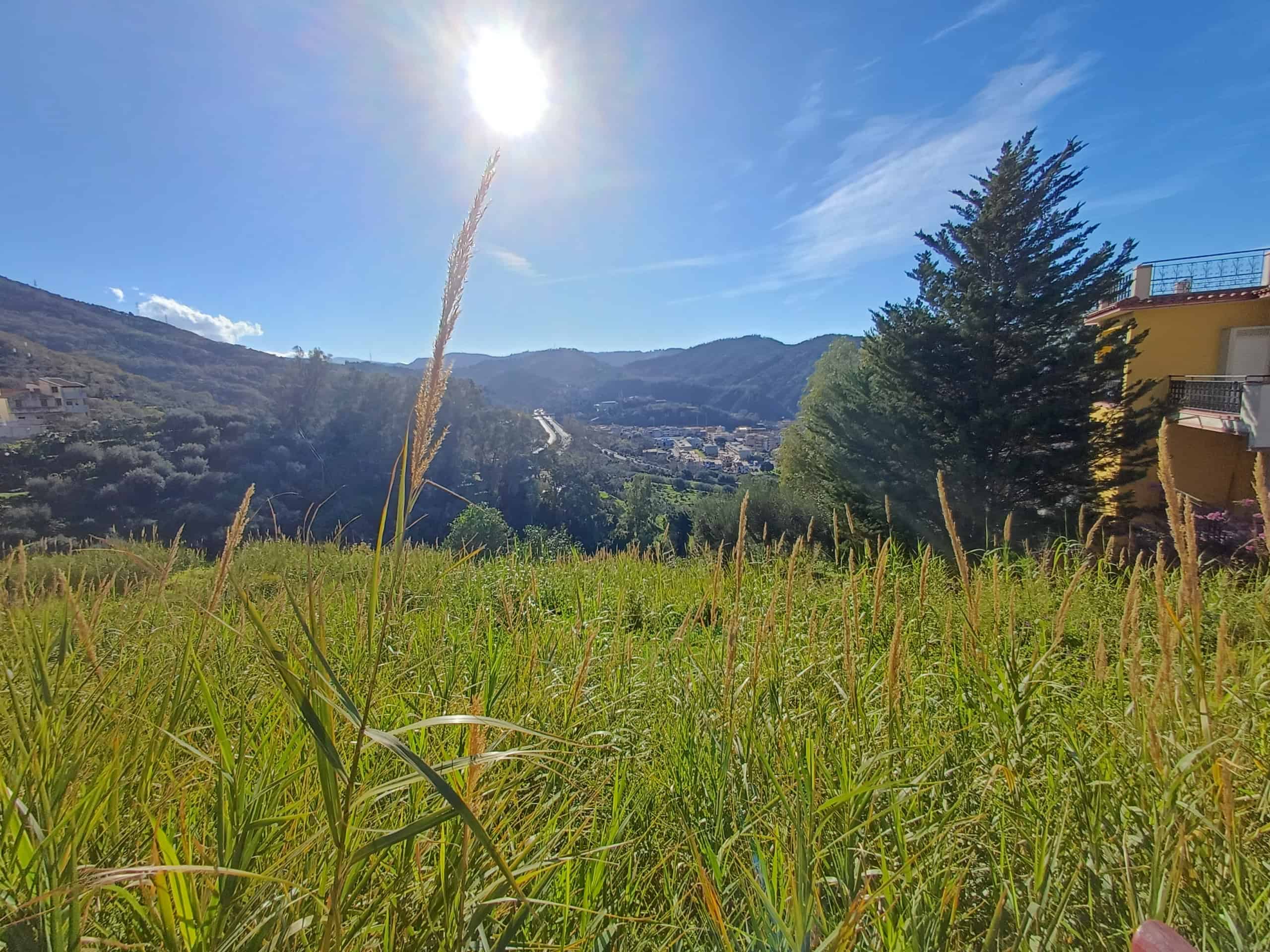 Prato verde e soleggiato con montagne sullo sfondo, cielo azzurro e sole brillante, vista panoramica di un paesaggio naturale e tranquillo in Italia.