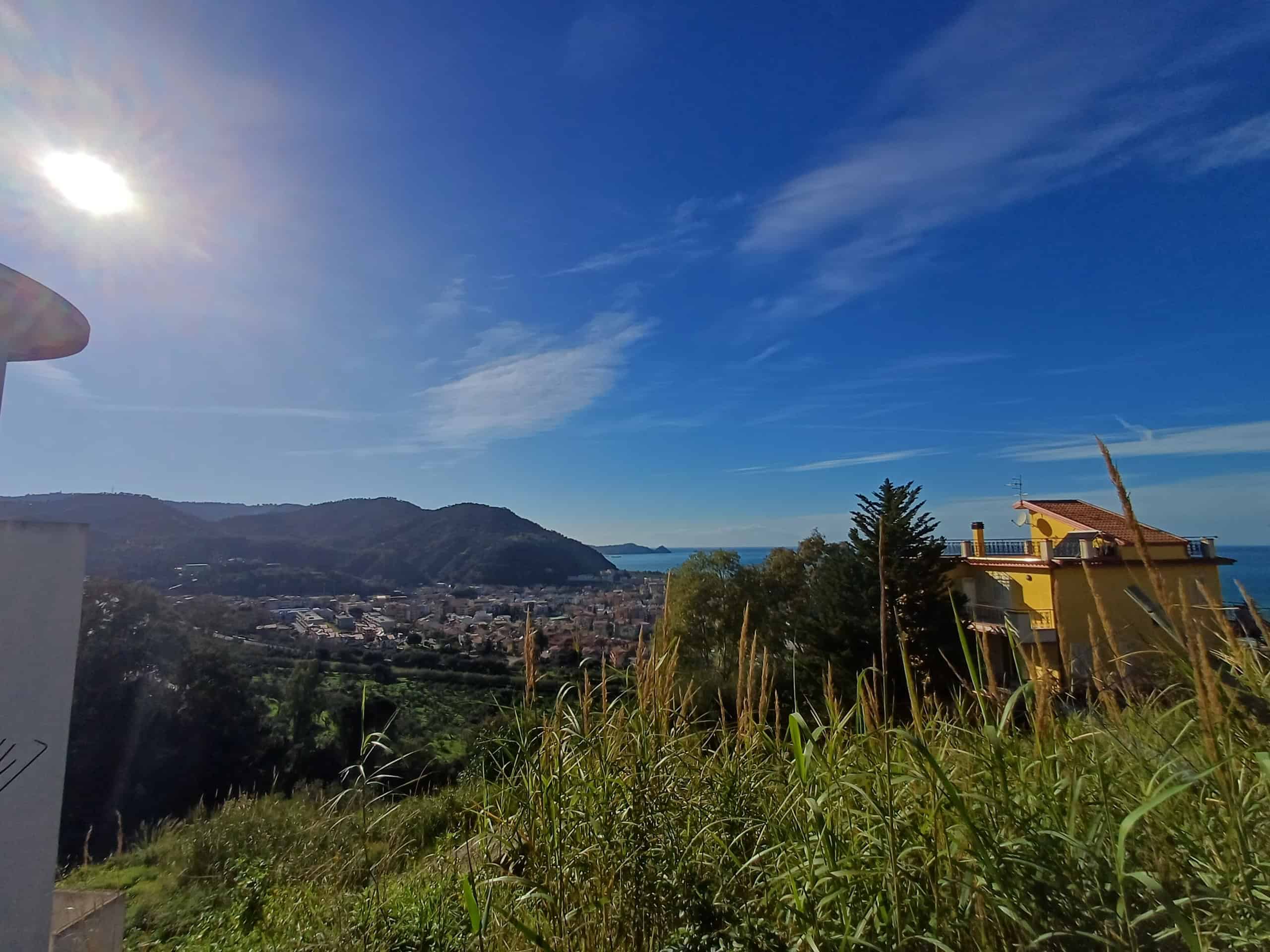 VISTA PANORAMICA di un paesaggio collinare con città, mare e cielo azzurro, con edifici e vegetazione, sotto un cielo sereno con nuvole leggere e sole splendente.