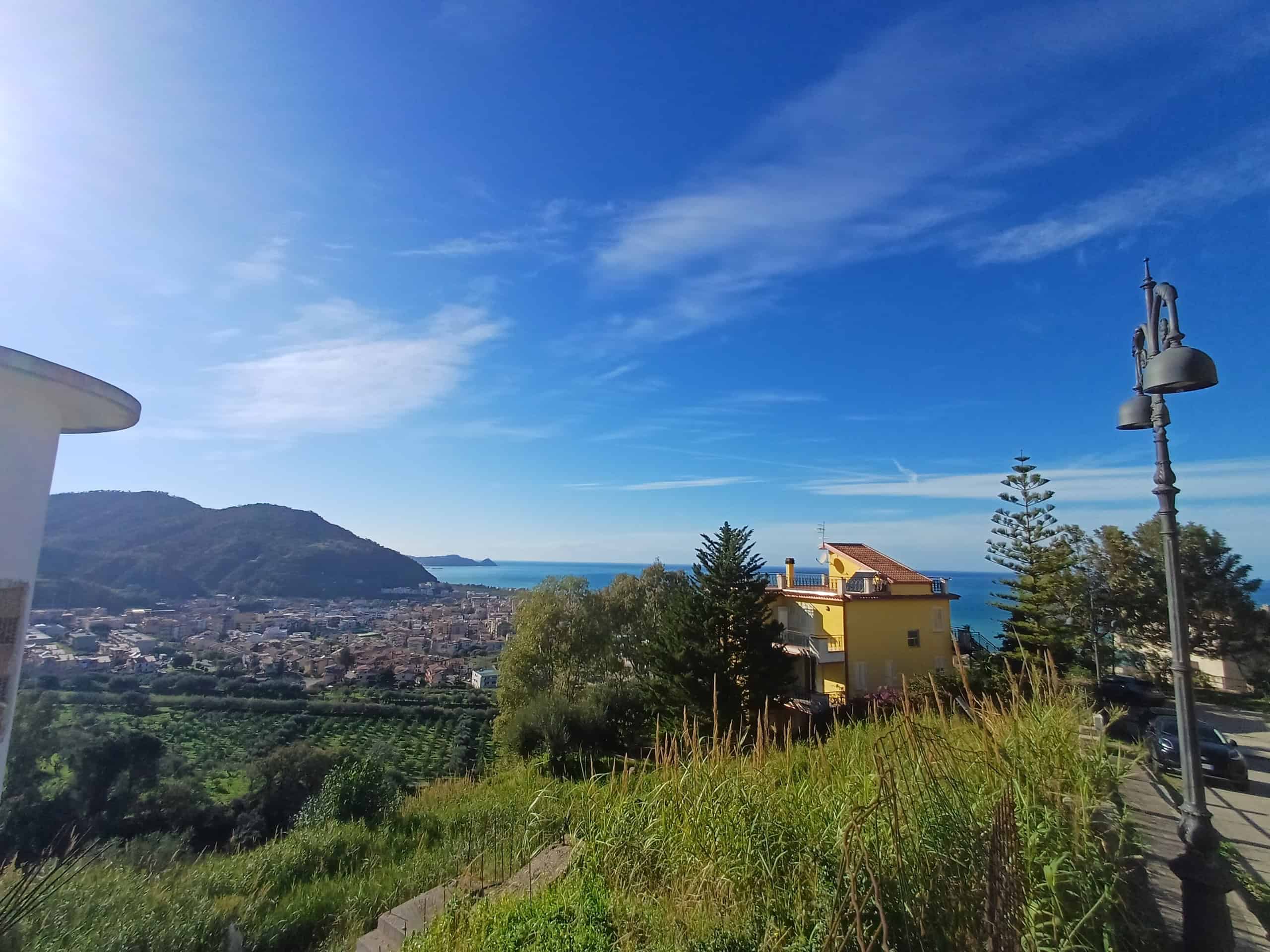 Verde paesaggio collinare con vista mare, case colorate e lampioni lungo strada, cielo azzurro con nuvole leggere, scenario tranquillo e suggestivo, ideale per immobili di lusso e residenze panoramich.