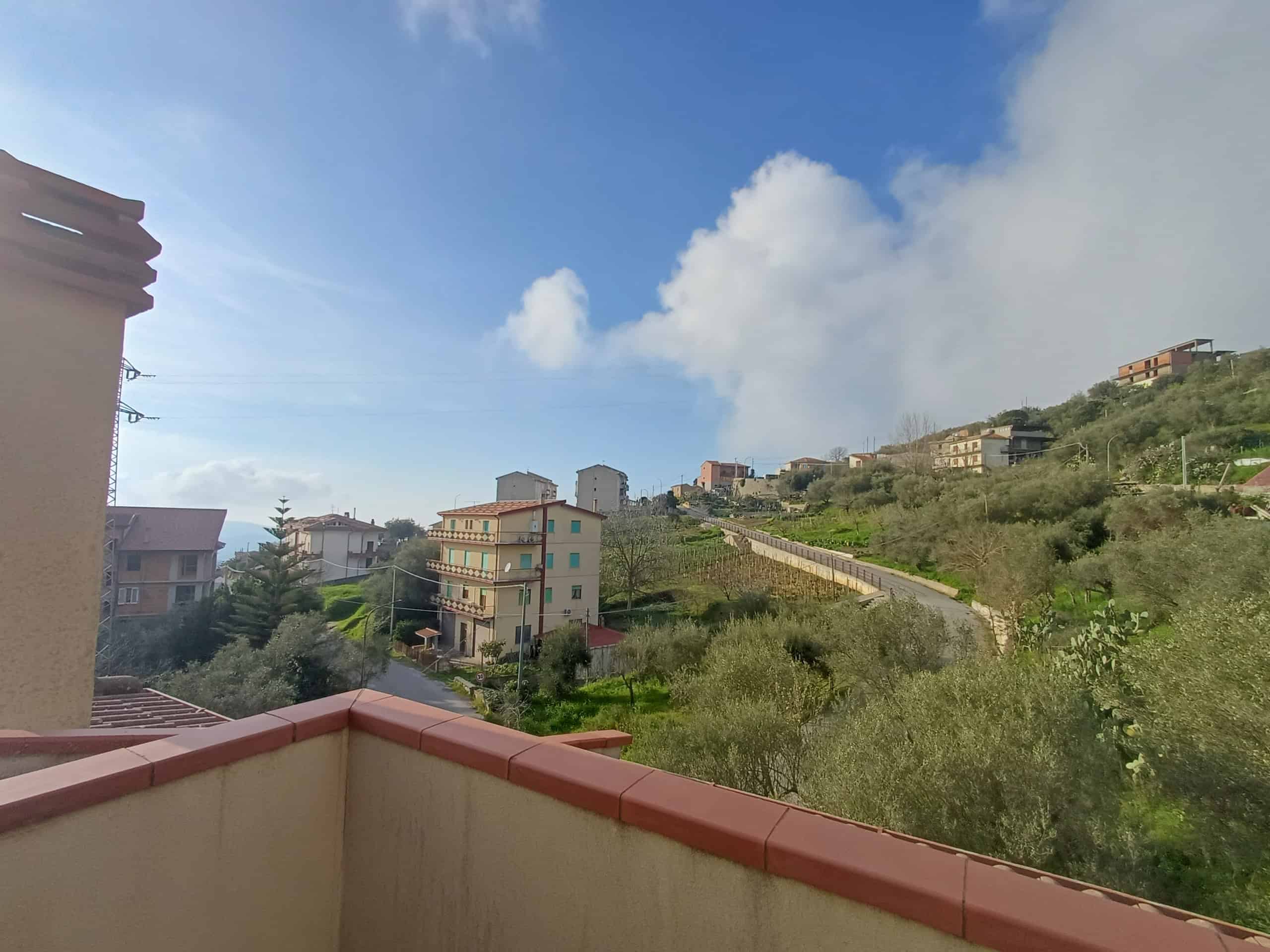 Vista panoramica di Ficarra con case e colline verdi sotto un cielo blu con nuvole bianche, visibile dalla terrazza di una casa indipendente.