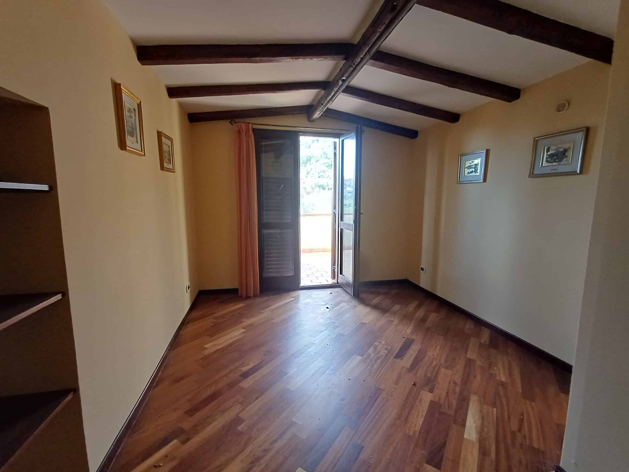Interno di una camera da letto con pareti beige, parquet in legno e soffitto con travi a vista, con accesso a un balcone esterno.