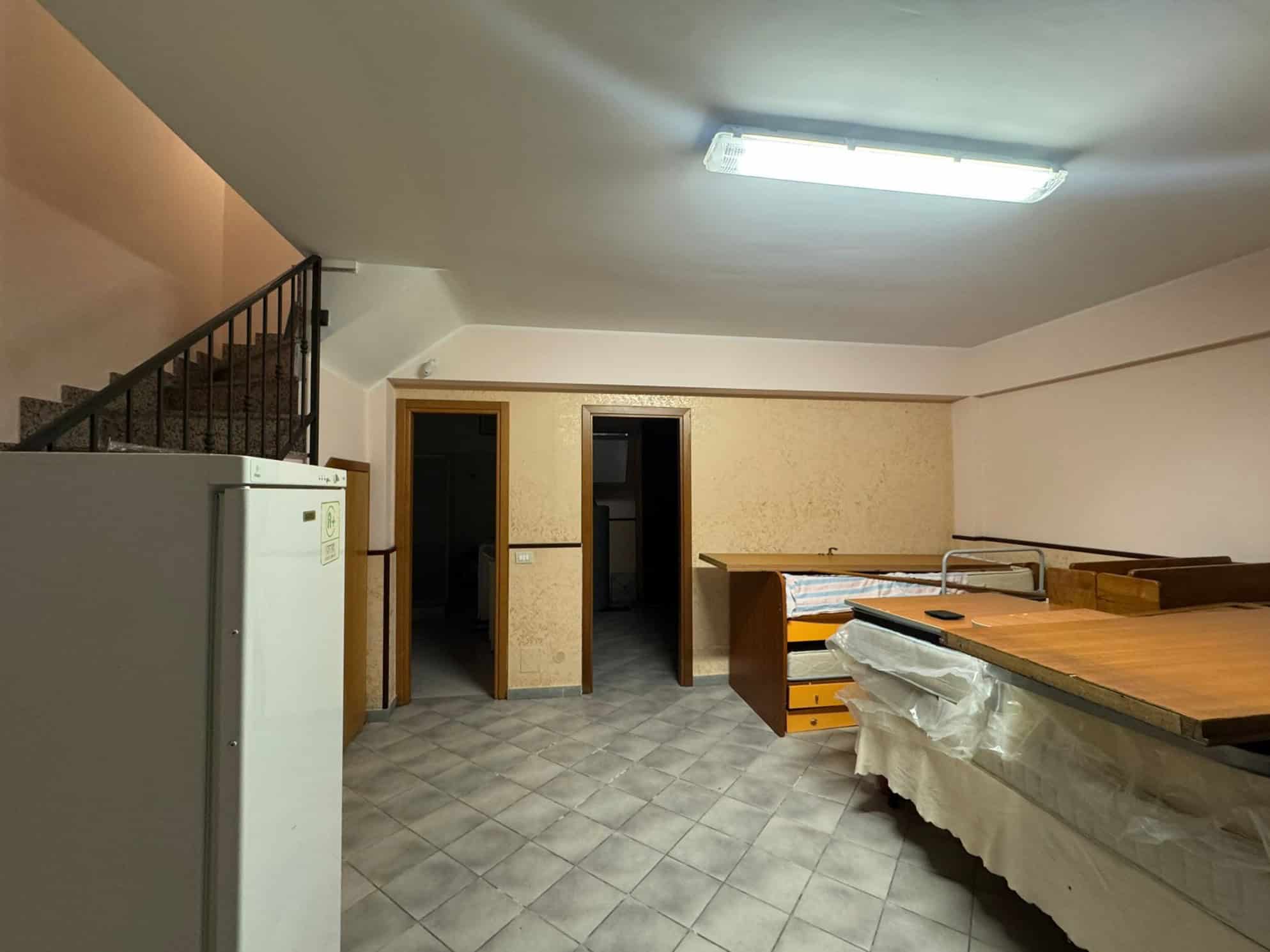 Spazio interno di un appartamento con pareti beige, pavimento in piastrelle e arredi in legno, illuminato da luce fluorescente sul soffitto, con porte che conducono ad altre stanze e un frigorifero bi.