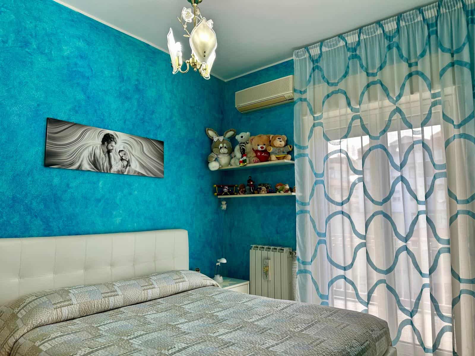 Un letto matrimoniale in una camera da letto moderna con pareti blu vivaci, decorazioni di peluche su una mensola e tende leggere con motivo geometrico, aria condizionata e radiatore visibili.