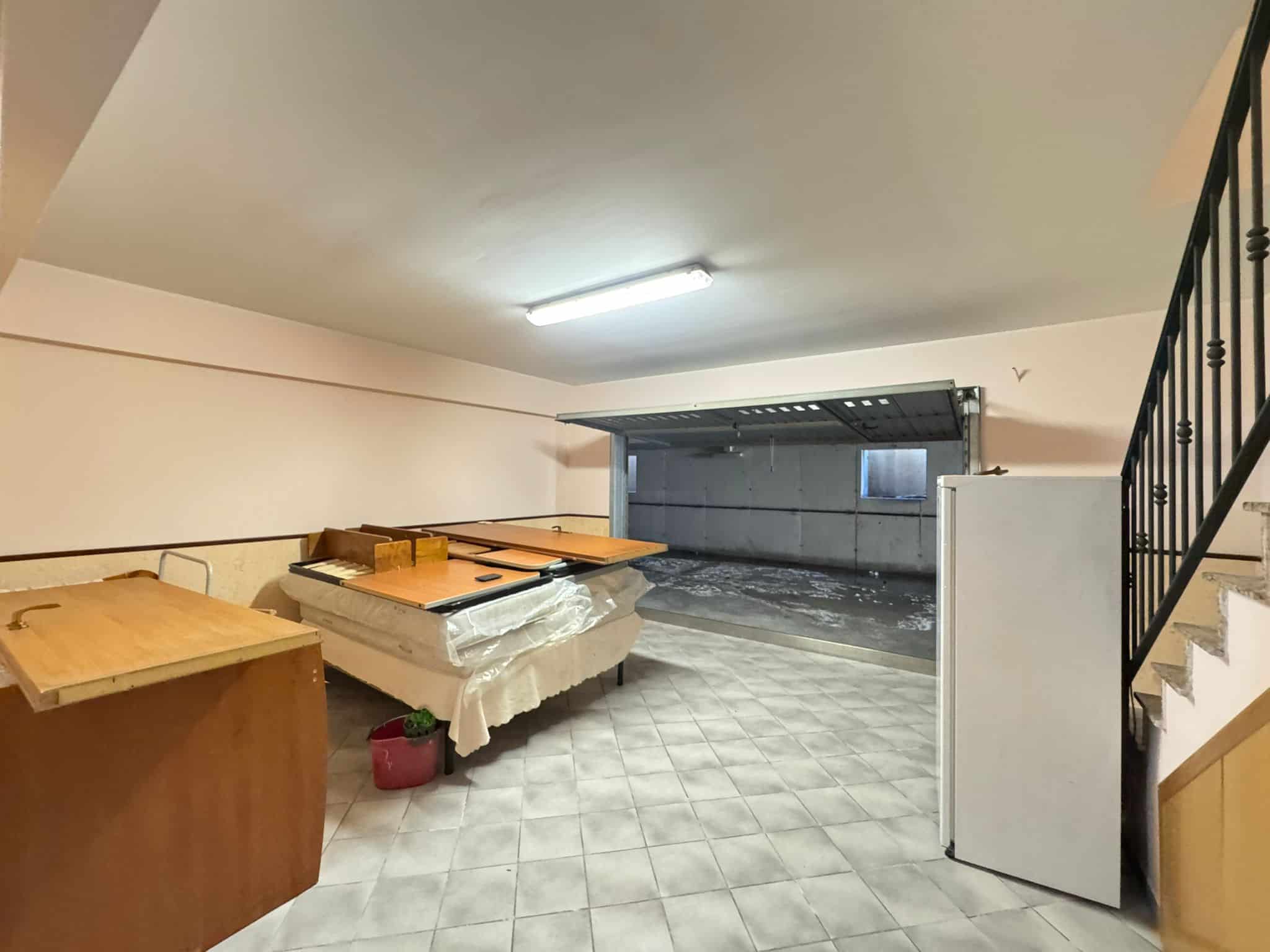 Ampio spazio commerciale o magazzino con pavimento in piastrelle, pareti bianche e illuminazione a LED, con ingresso indipendente e scala che conduce a un piano superiore, ideale per attività di vendi.