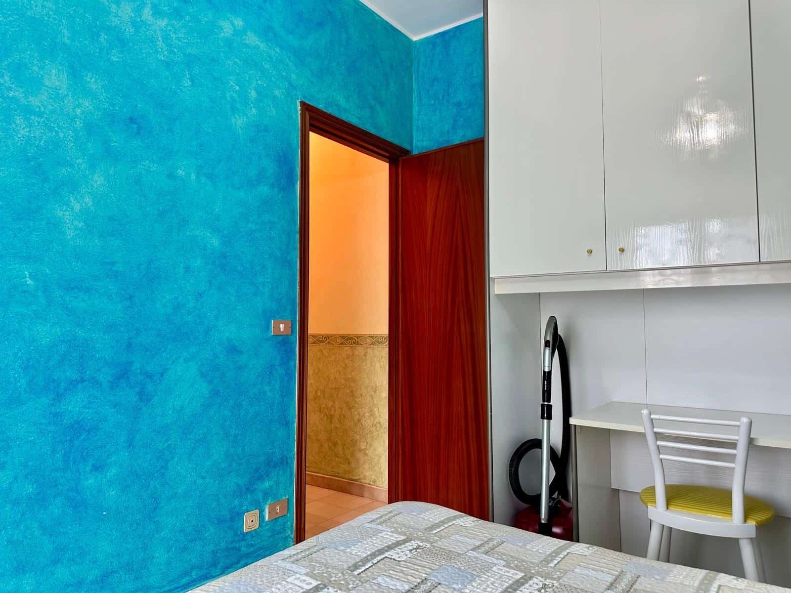 Soggiorno con pareti blu vivaci, armadio bianco e angolo studio con sedia gialla, ideale per ambienti moderni e funzionali in appartamenti in vendita o affitto a NextCasa.