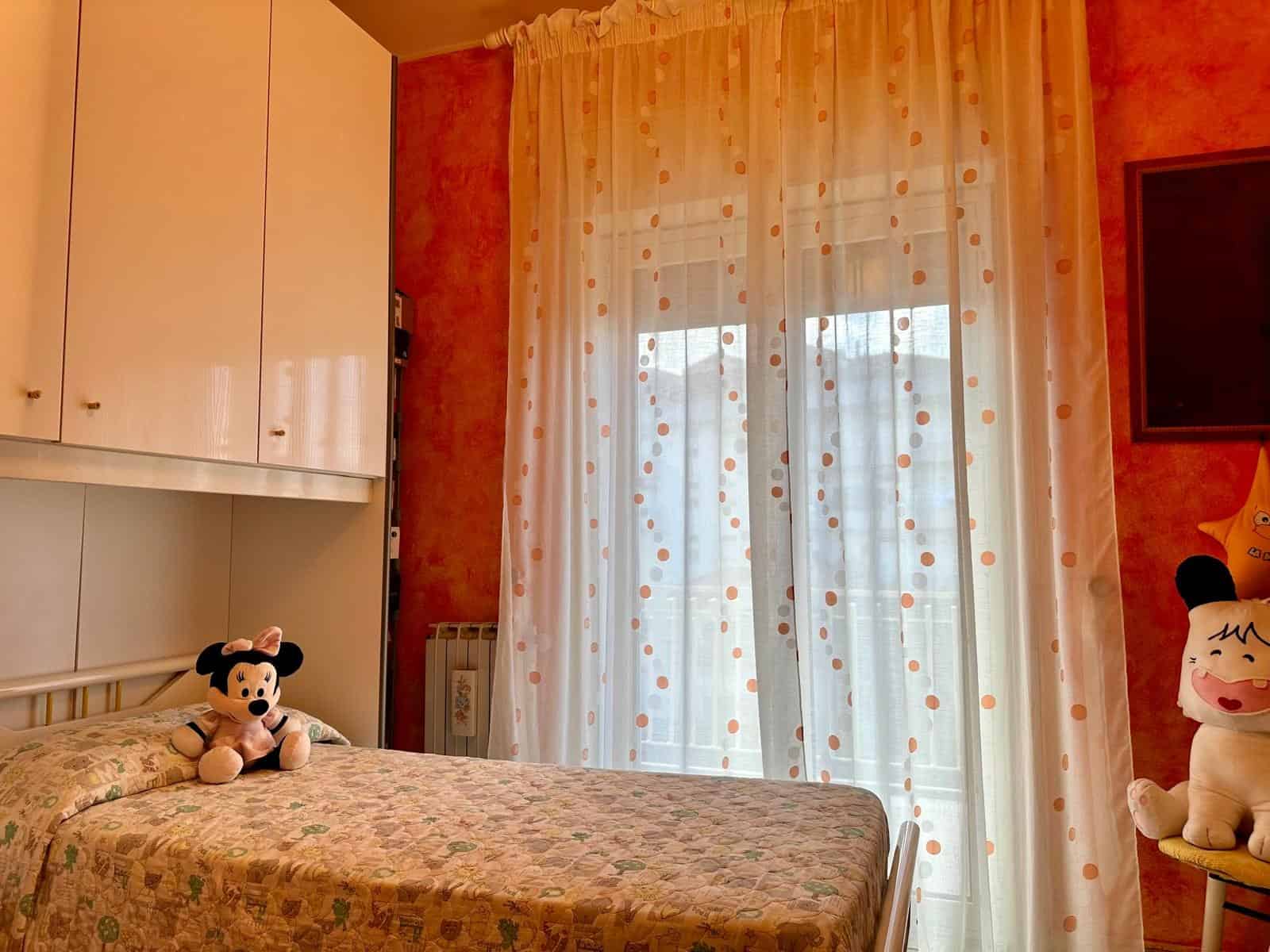 Mickey Mouse e Minnie Mouse in una stanza da letto con tende trasparenti e pareti arancioni, arredamento accogliente e decorazioni per bambini, ambiente luminoso e familiare.