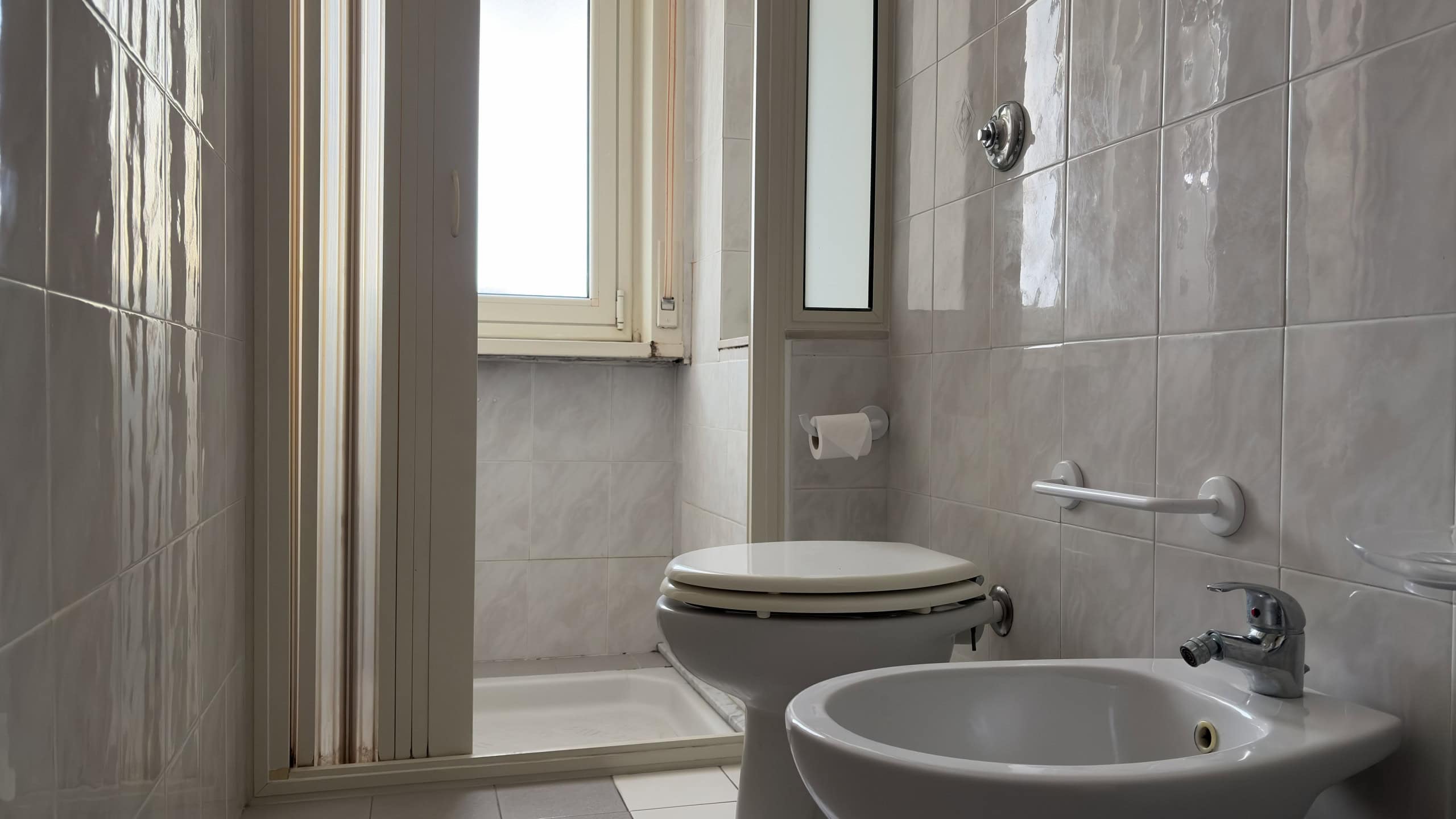 Lavabo e servizi igienici in bagno con pareti rivestite in piastrelle beige, finestra per ventilazione naturale, accessori di sicurezza e comfort.