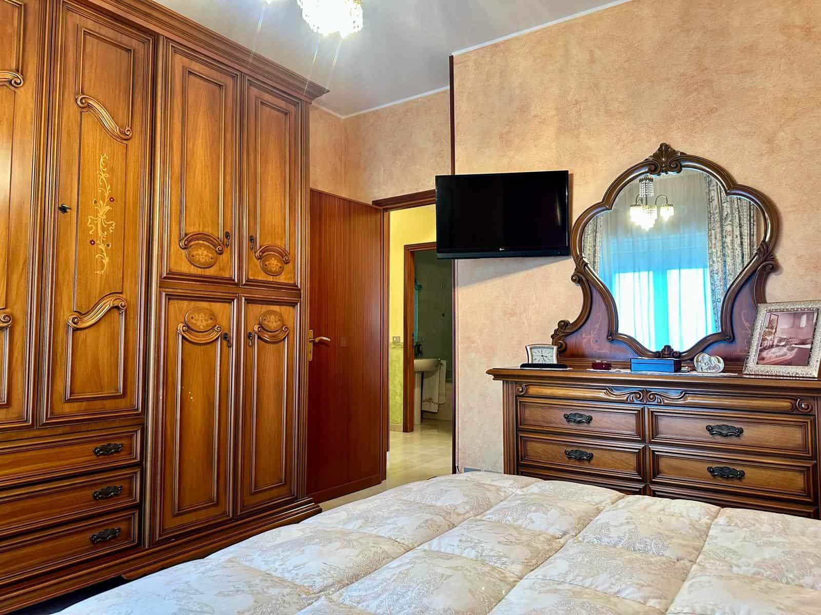 Camera da letto con armadio in legno, specchio e TV, arredamento classico, ambiente accogliente e ben illuminato.