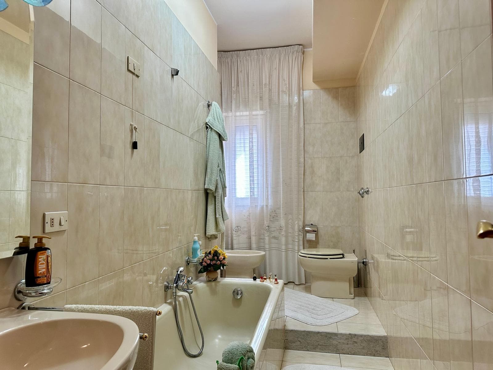 Lavabo e vasca da bagno in un bagno moderno con pareti rivestite in piastrelle beige, asciugamani appesi e finestra con tende leggere, ambiente luminoso e accogliente.