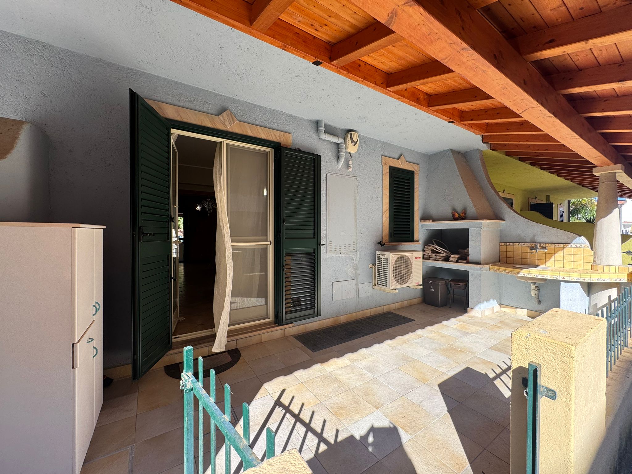 Porte finestra con persiane verdi che si affaccia su un balcone soleggiato con arredi esterni e vista sul mare, ideale per relax e convivialità all'aperto.