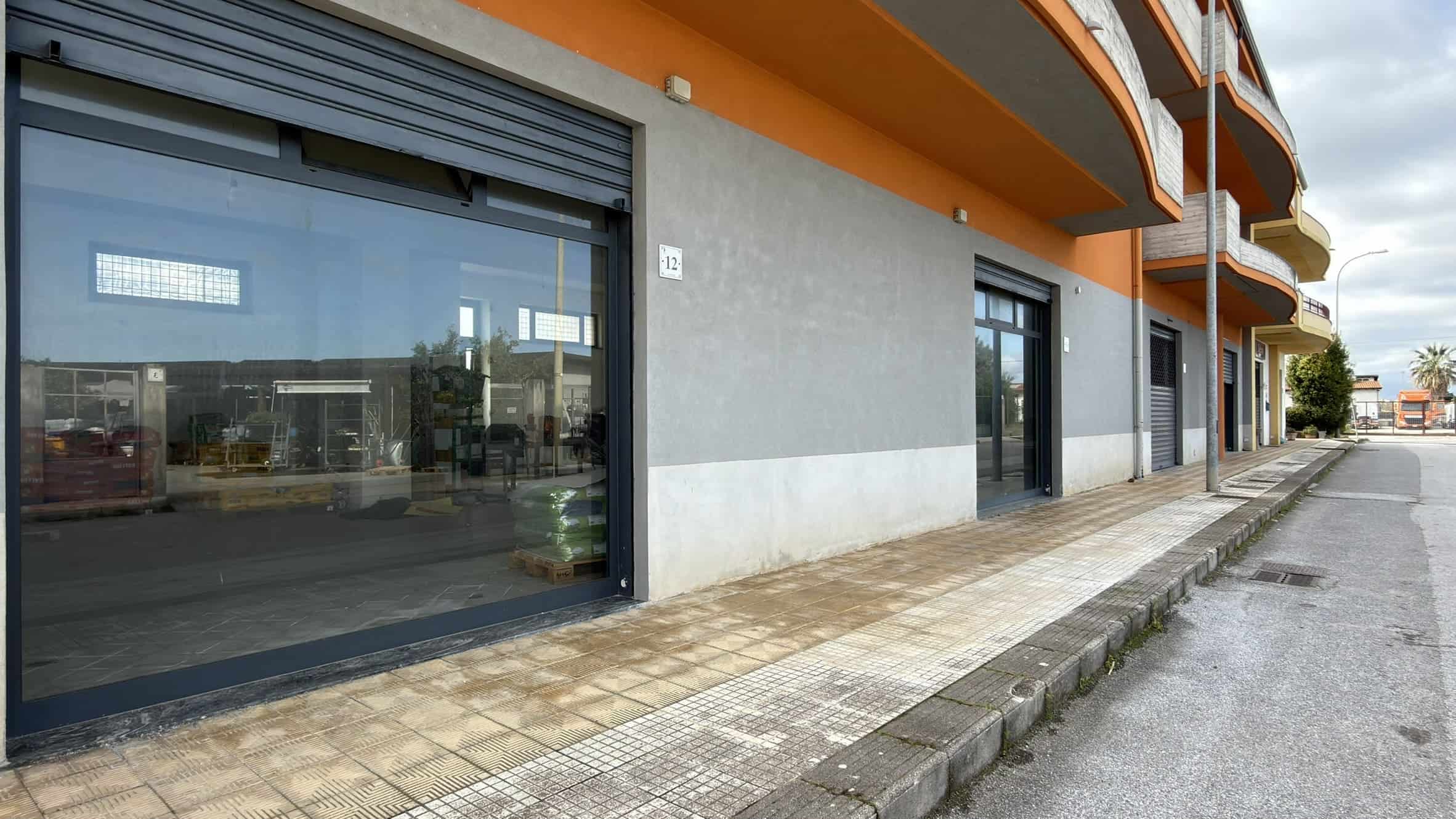 Vetrine commerciali con grandi finestre e porte scorrevoli in un edificio moderno, situato lungo un marciapiede in un'area urbana, con balconi sovrastanti e un cielo nuvoloso sullo sfondo.