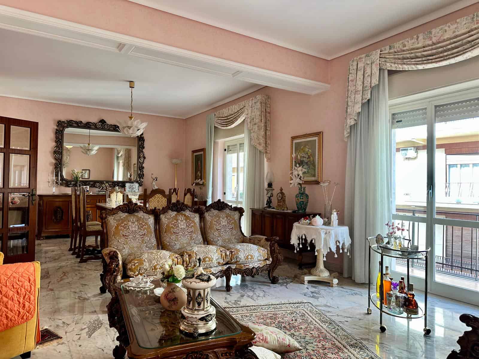 Interno di un elegante appartamento con arredi classici, divani in stile barocco e ampie finestre con tende floreali, luminoso e accogliente ambiente residenziale.