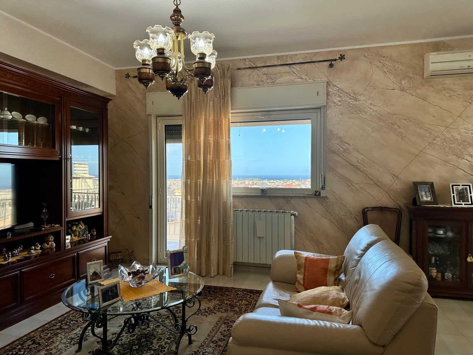 Ampia vista sulla città attraverso la finestra del soggiorno, arredato con divano beige, tavolino in vetro e mobile in legno, illuminato da lampadario antico, ideale per ambienti residenziali di lusso.