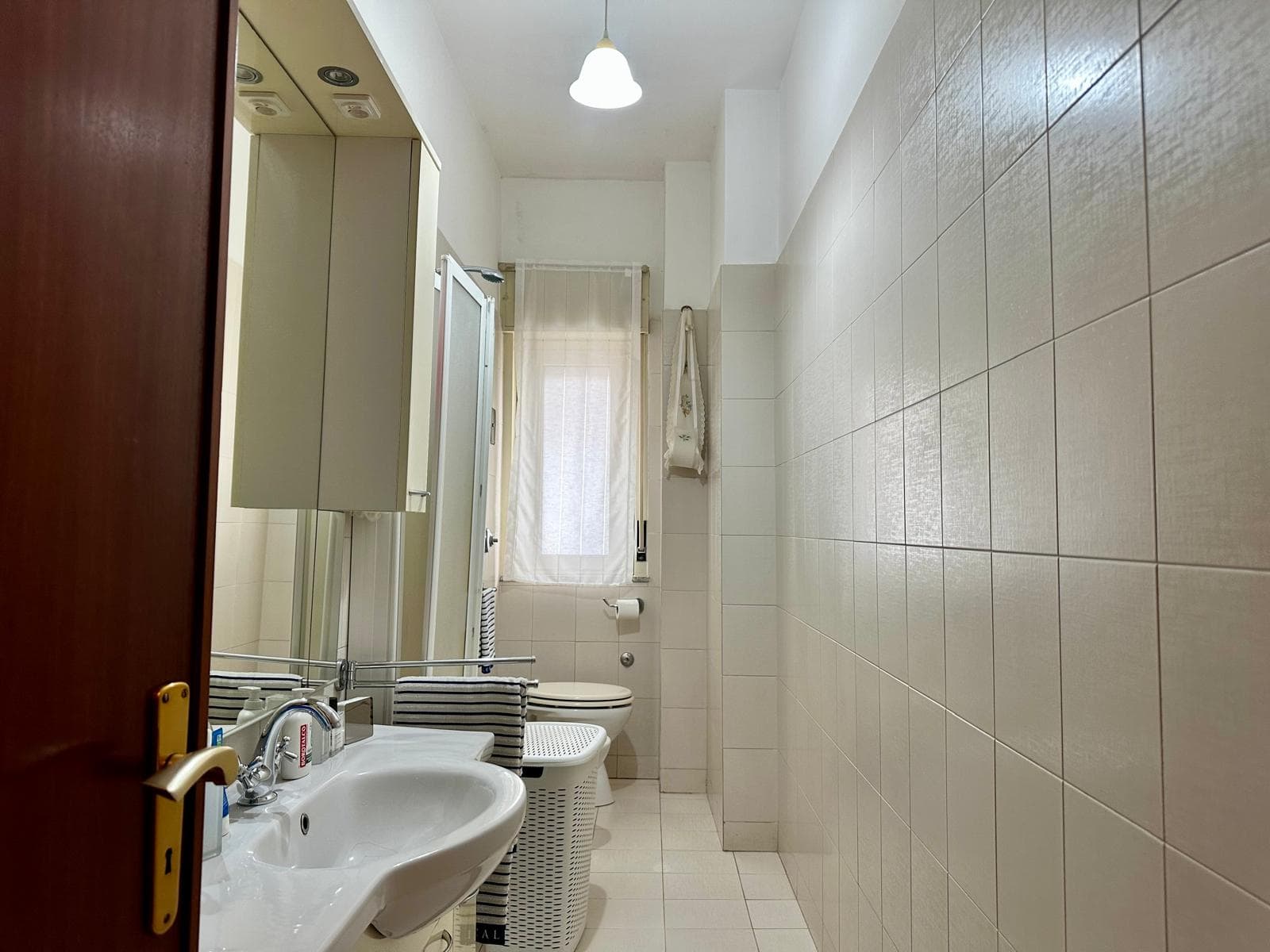Lavabo e servizi igienici moderni in un bagno luminoso con pareti rivestite di piastrelle chiare e finestra con tende leggere, ideale per appartamenti in vendita o affitto a NextCasa.