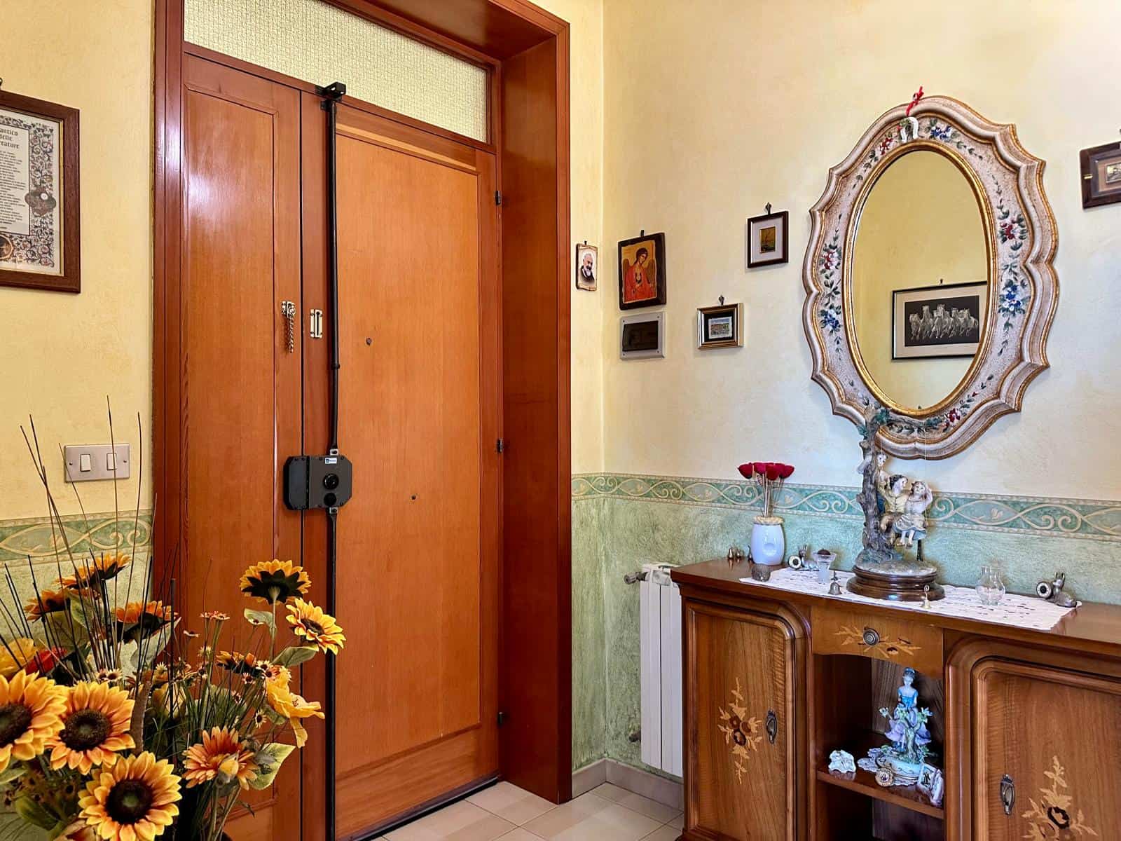 Porta blindata in legno con serratura di sicurezza, decorata con dettagli eleganti, in un ingresso accogliente con specchio decorativo, mobili in stile classico, e fiori freschi, creando un ambiente c.