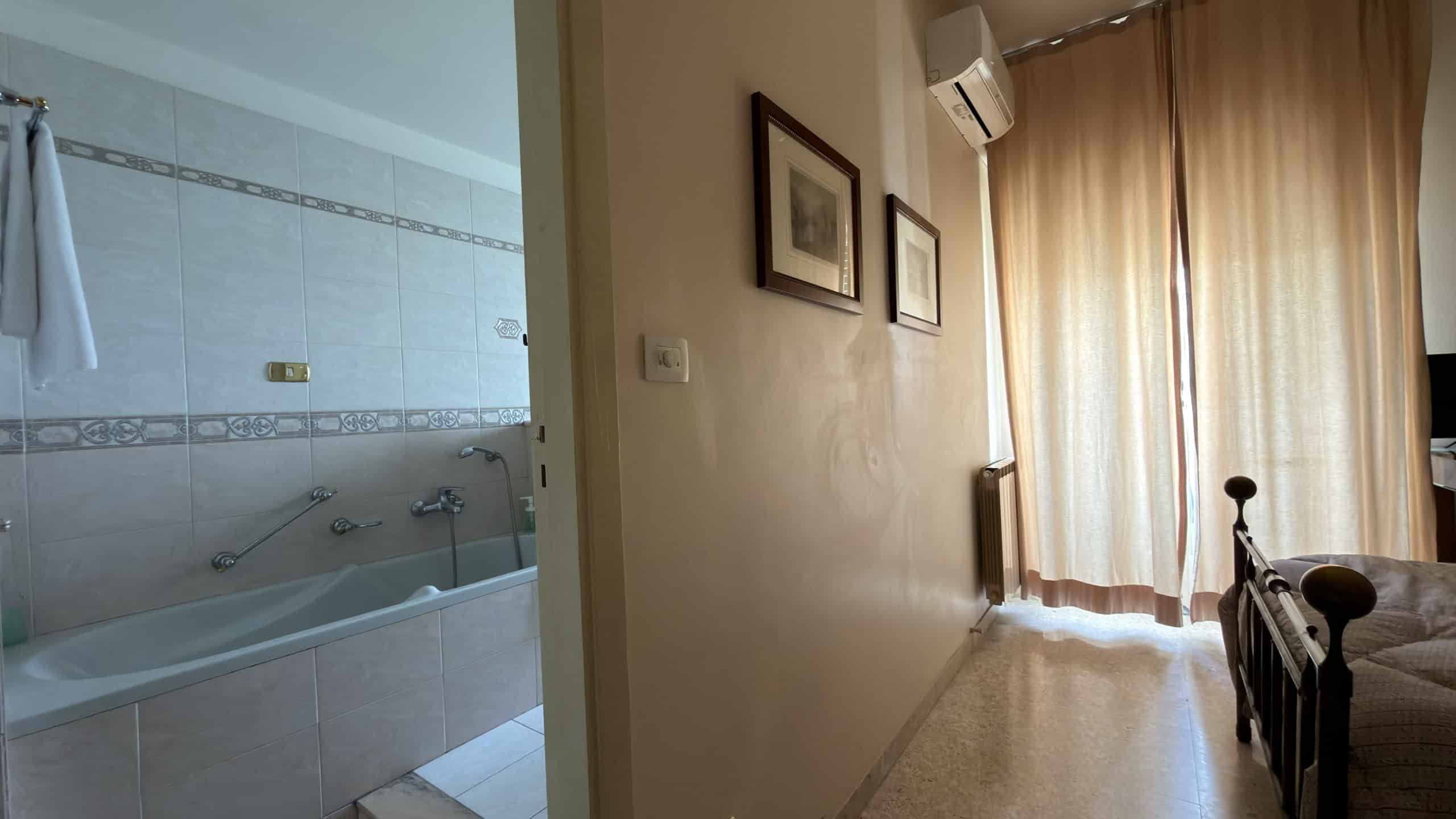 Ampia vista di un soggiorno con pareti beige, decorato con quadri e dotato di aria condizionata, con accesso a una camera da letto e a un balcone con tende leggere, ideale per affitti turistici o resi.