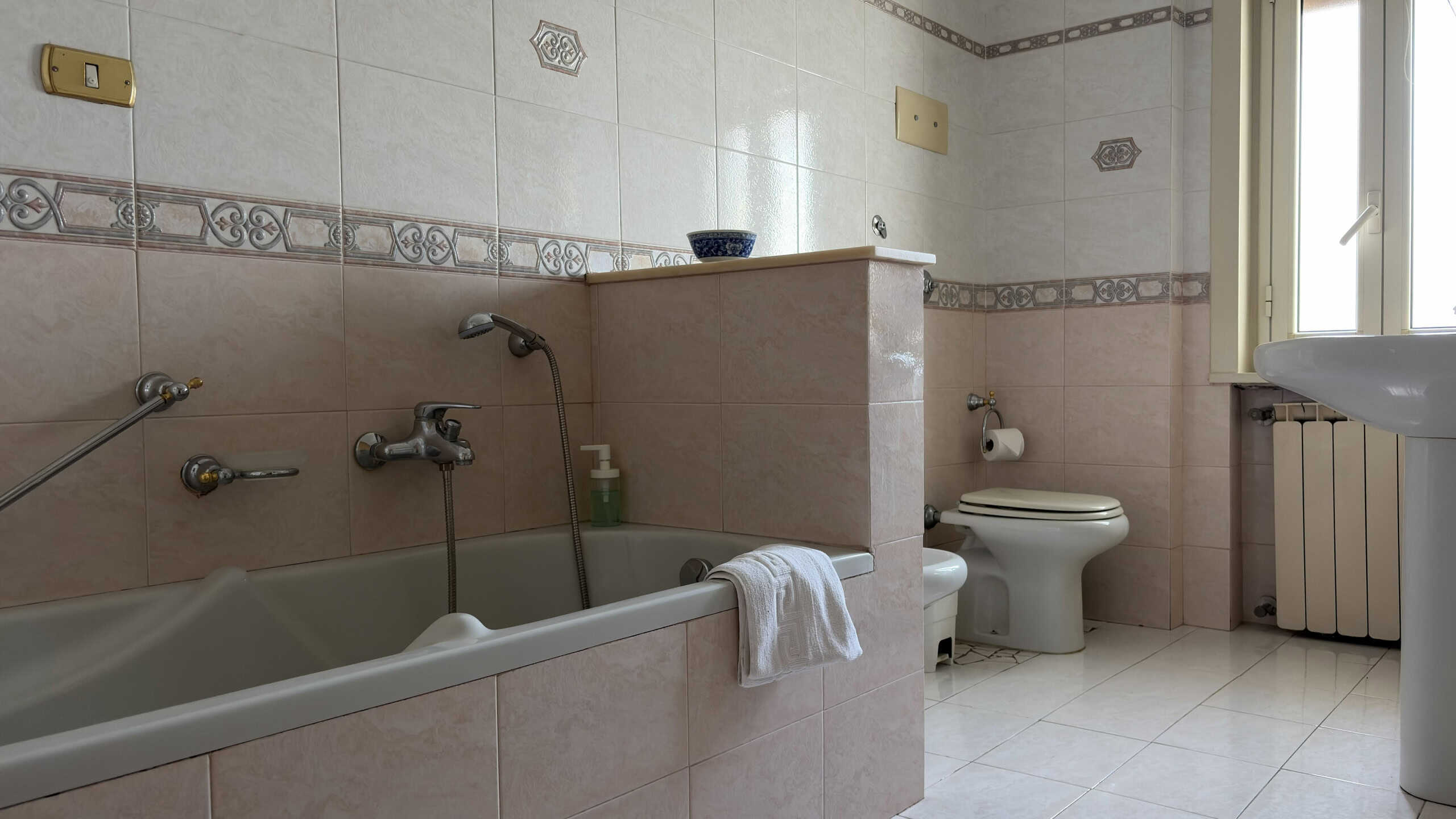 Lavabo e vasca da bagno in un bagno spazioso con piastrelle beige e finestra luminosa, ideale per ristrutturazioni e arredi moderni.