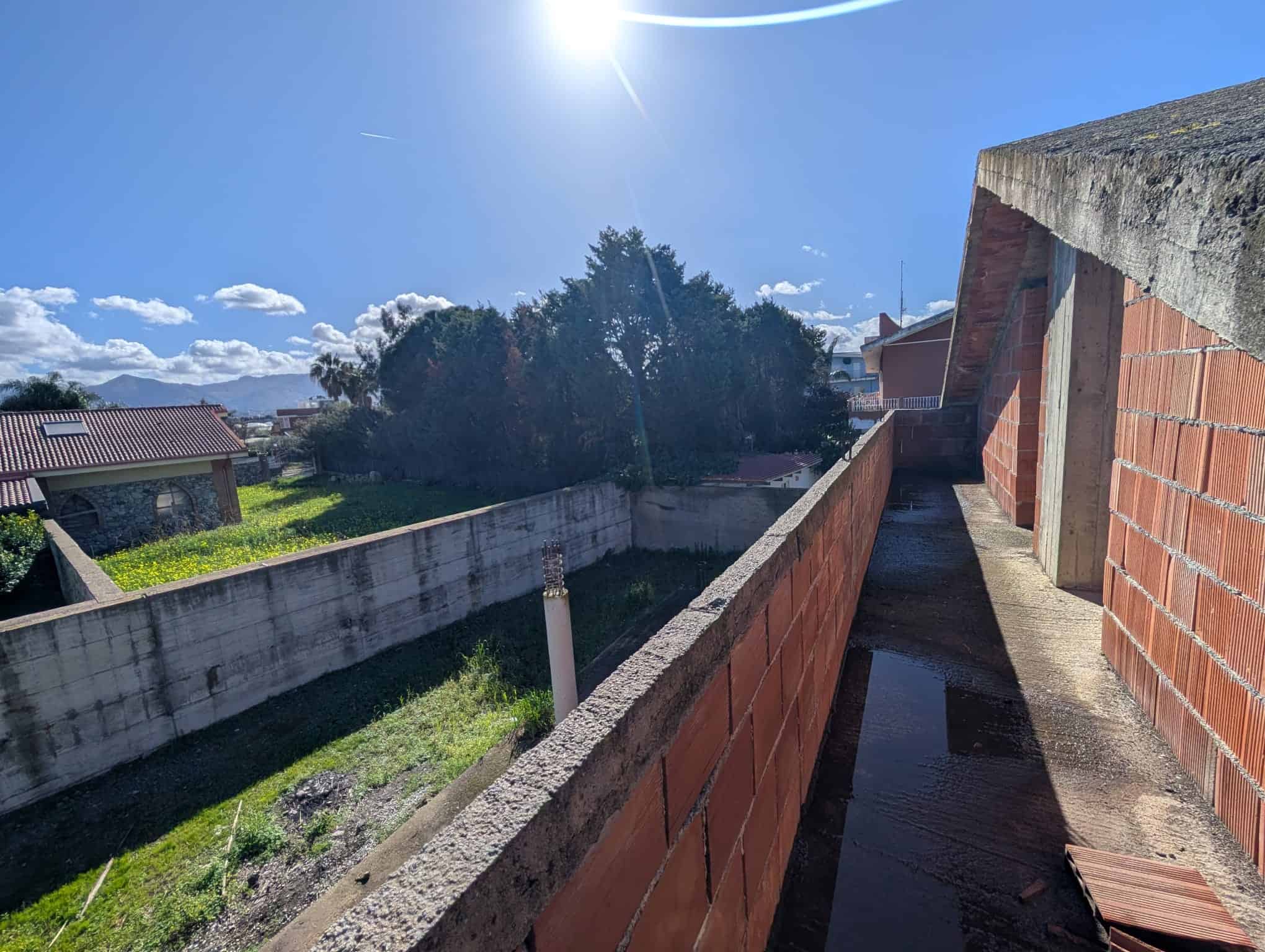 Finestra di un edificio in costruzione con vista sul cielo sereno e sul paesaggio circostante, inclusi alberi e case, in un giorno soleggiato.