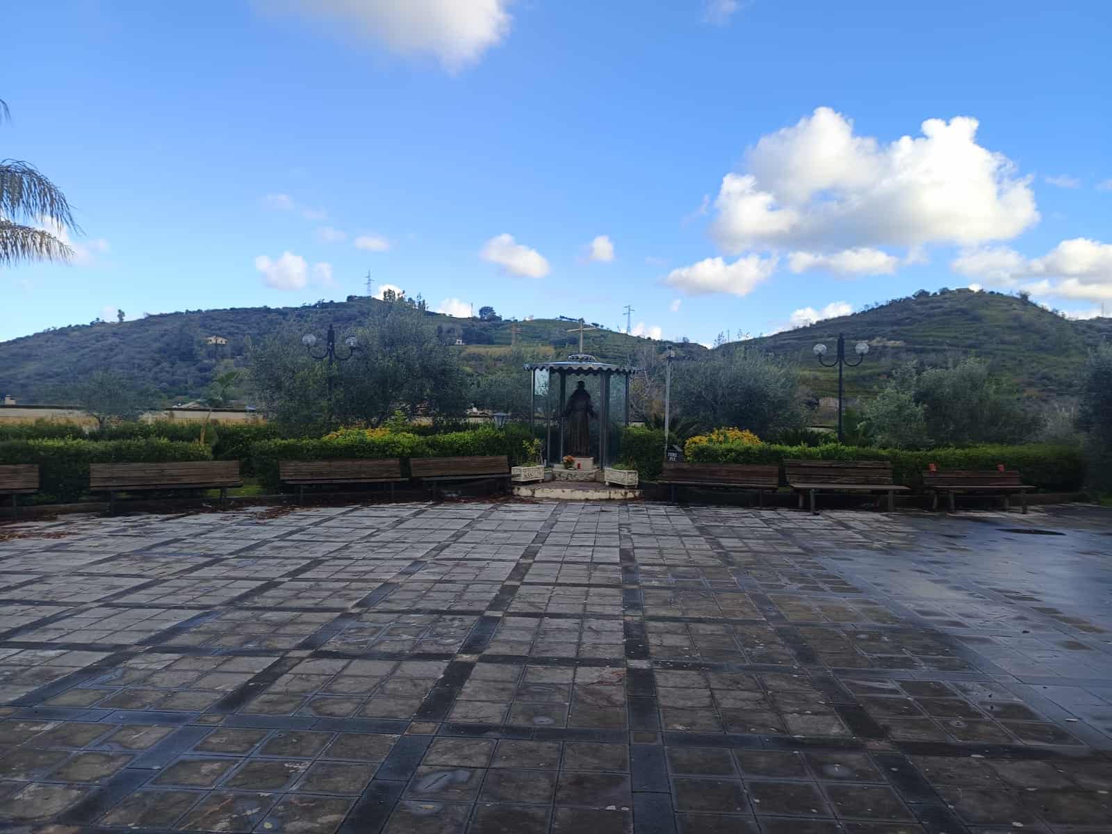 Un'area pubblica all'aperto con panchine, un gazebo e un paesaggio collinare sullo sfondo, sotto un cielo sereno con nuvole bianche, ideale per relax e socializzazione.