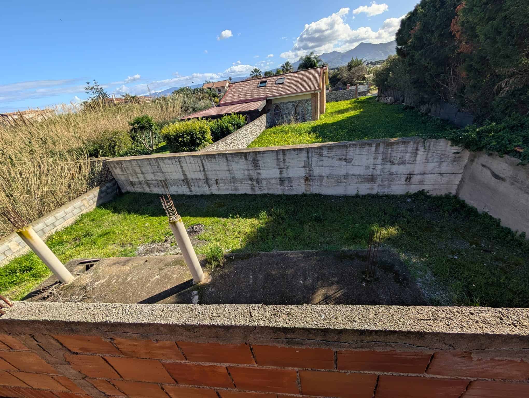 Vano scoperto con fondazione in cemento e recinzione in mattoni, pronto per la costruzione di una casa o un edificio residenziale, immerso in un'area verde con case e montagne sullo sfondo.
