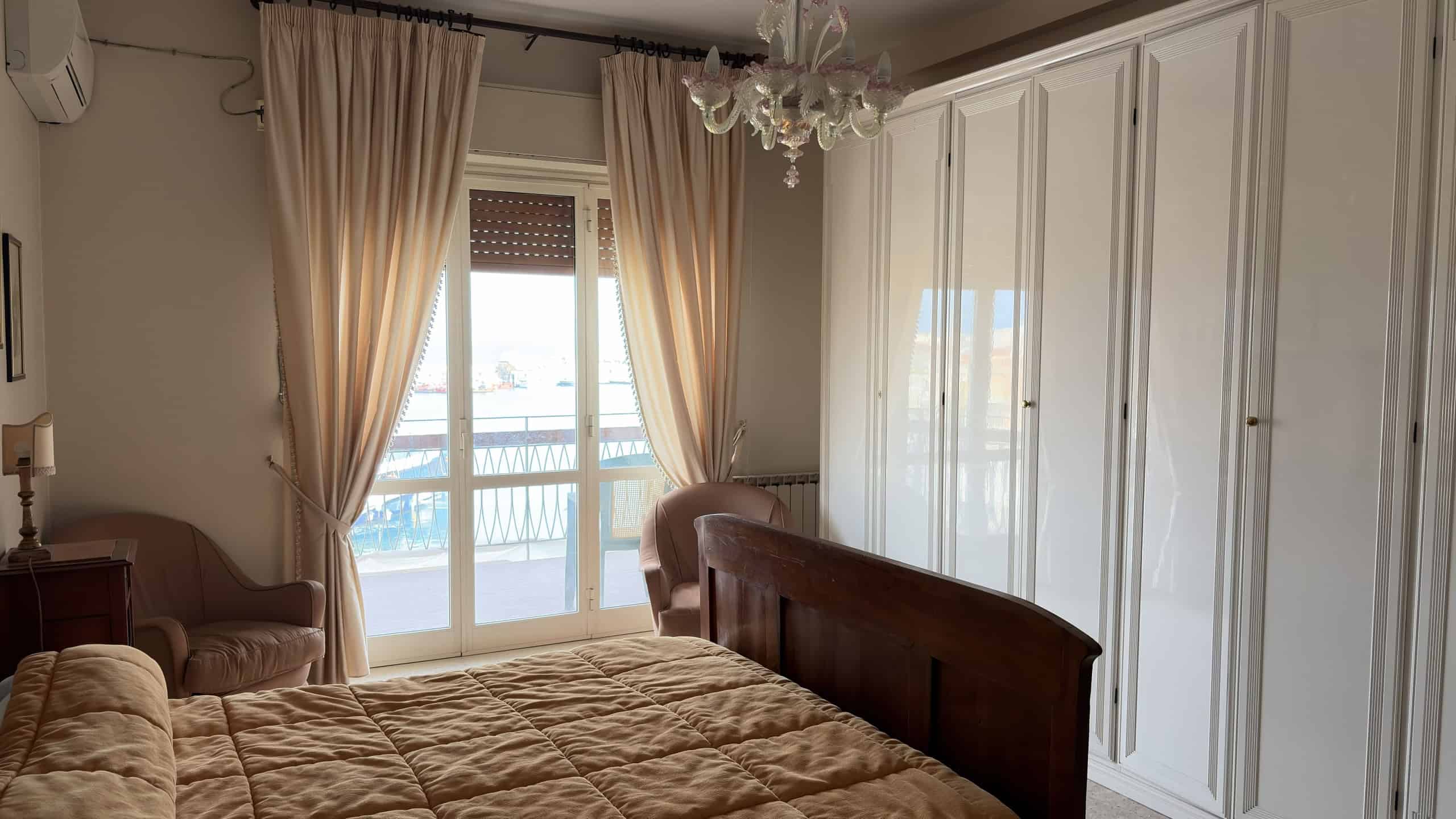 Letto matrimoniale in una camera da letto luminosa con grandi finestre e tende beige, arredi eleganti e armadi a muro bianchi, ambiente accogliente e spazioso.