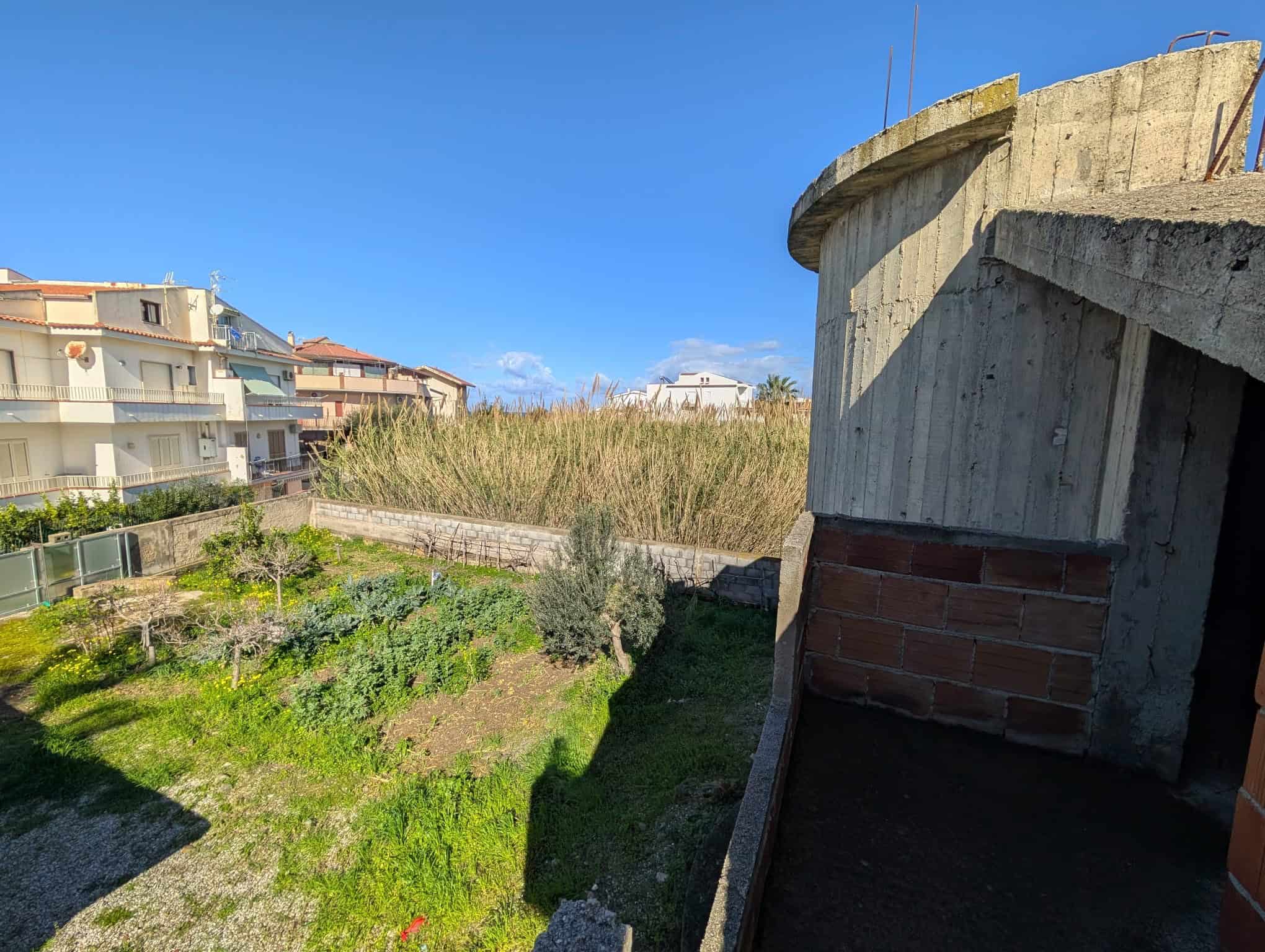 Pannello di costruzione in cemento e mattoni con vista su un giardino verde e case residenziali in lontananza, cielo azzurro e giornata soleggiata.